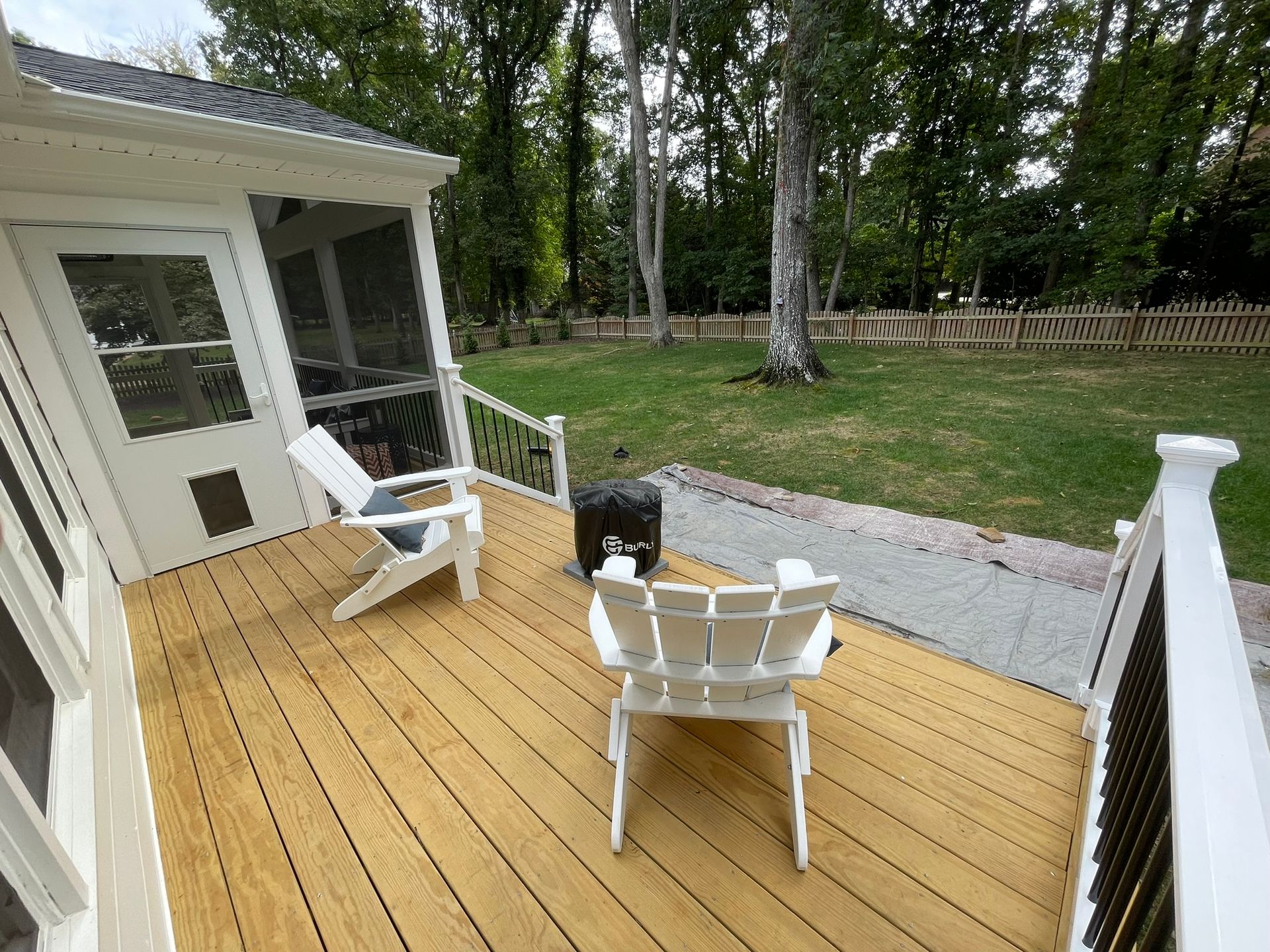 White patio chairs and a small table on a wooden deck beside a house, overlooking a grassy yard.