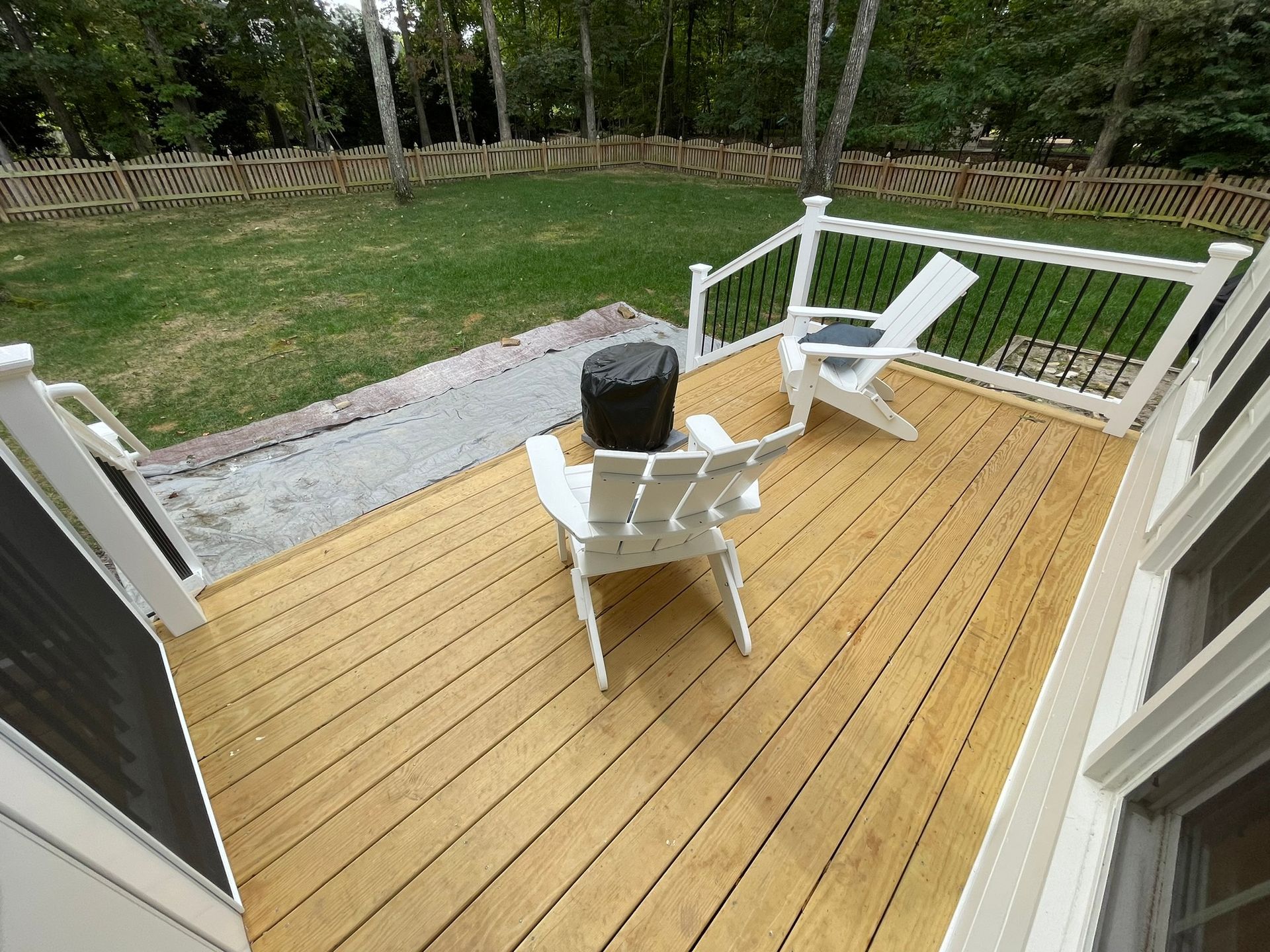 Wooden deck with two white chairs and a black planter, overlooking a grassy yard and trees.