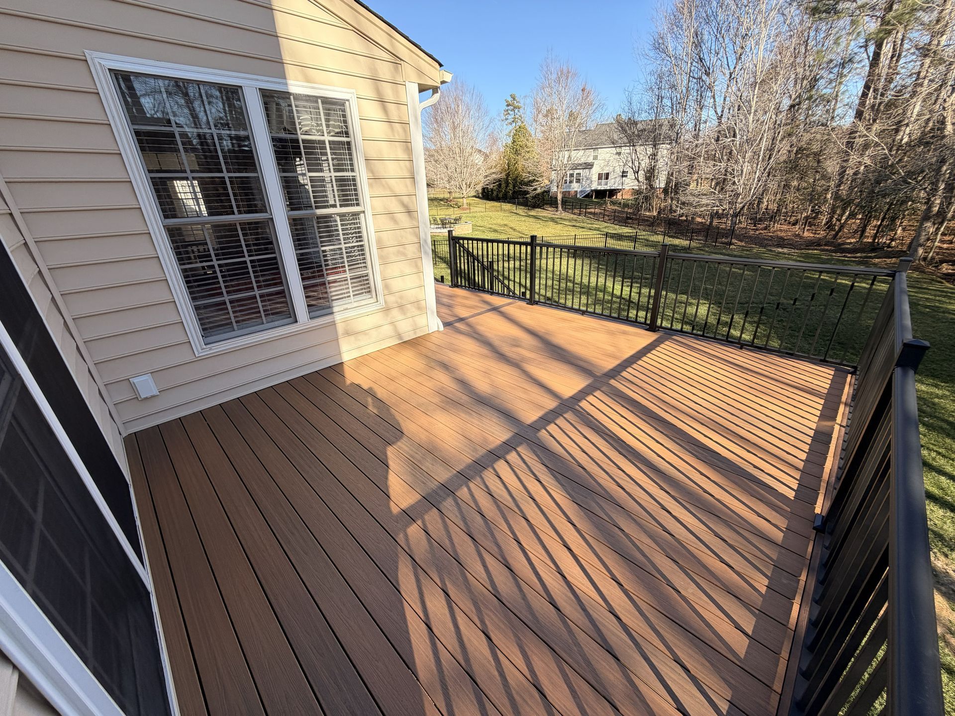 Wooden deck beside a beige house with white-trimmed windows and black railing, lit by long afternoon shadows.