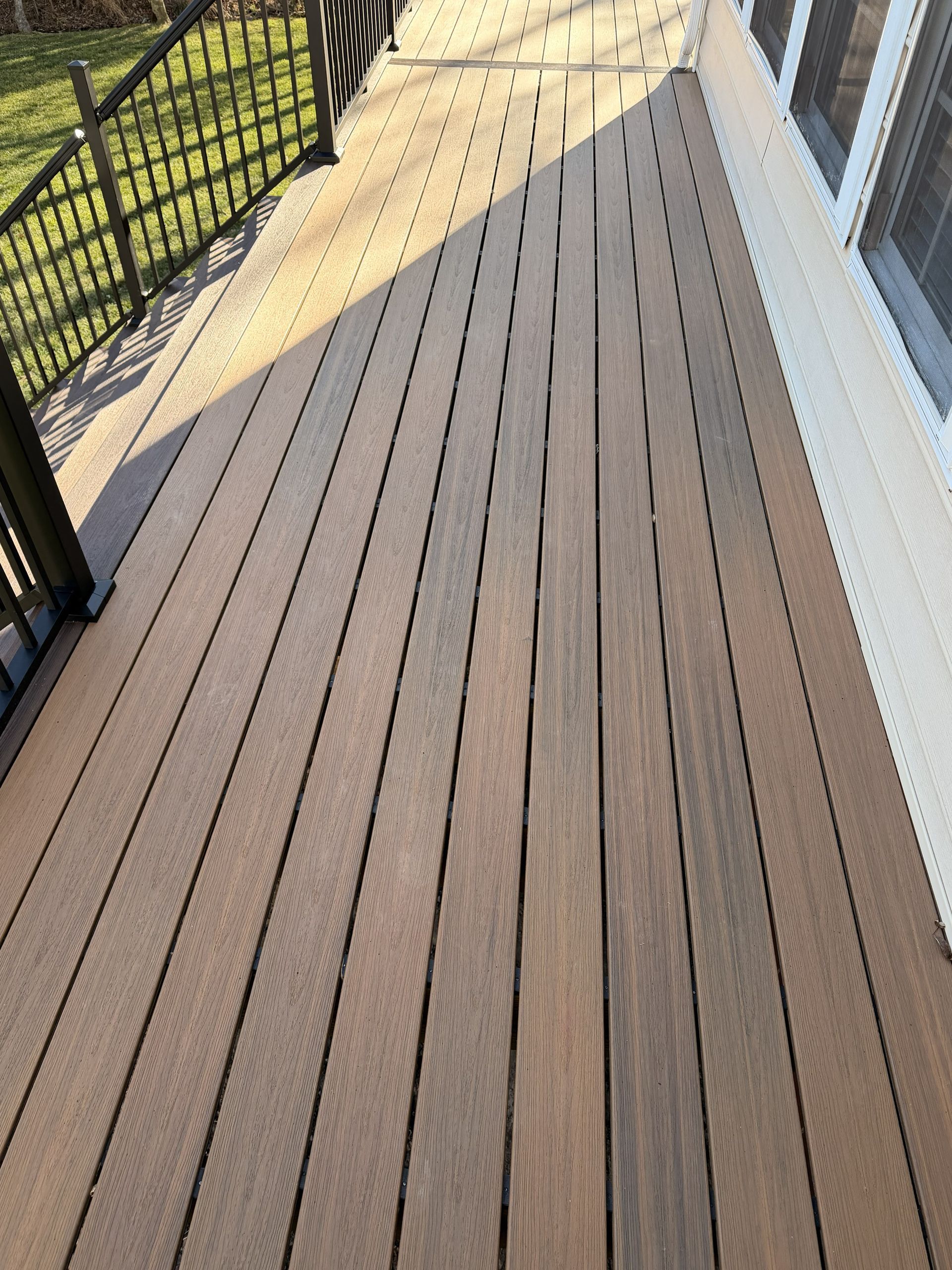 Wooden deck walkway beside a house, with a black railing and grass below.