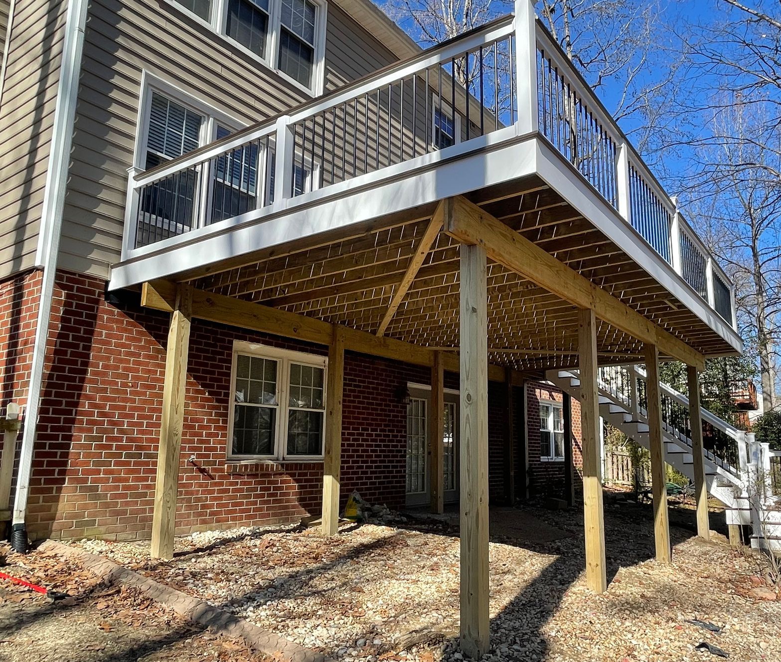 Raised wooden deck with white railing attached to a brick house, supported by posts over a gravel yard.