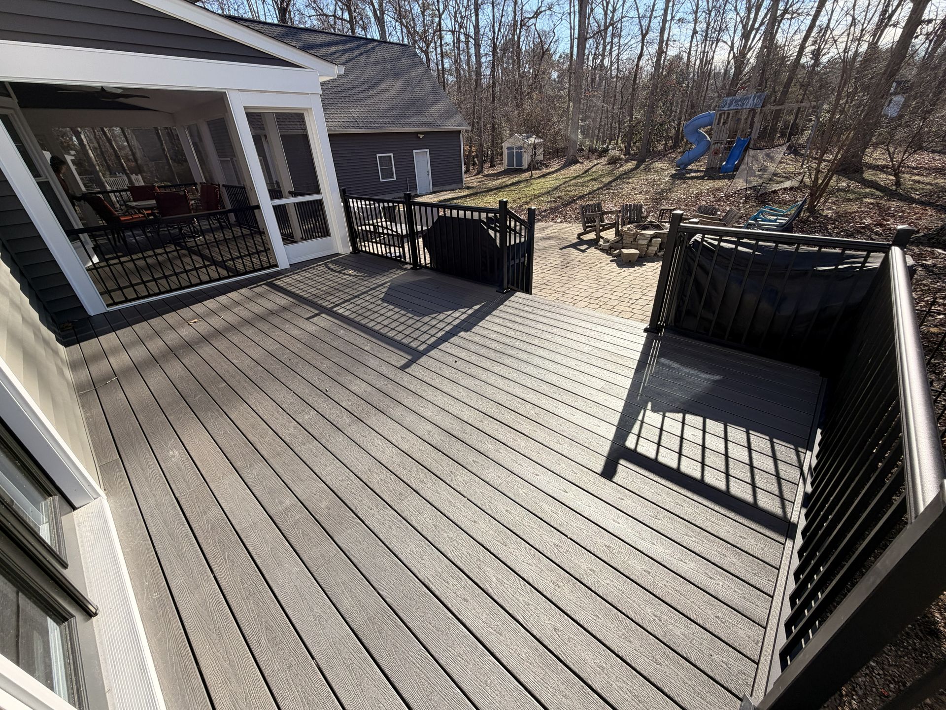 Spacious gray wooden deck with black railing and grill, overlooking a wooded backyard on a sunny day