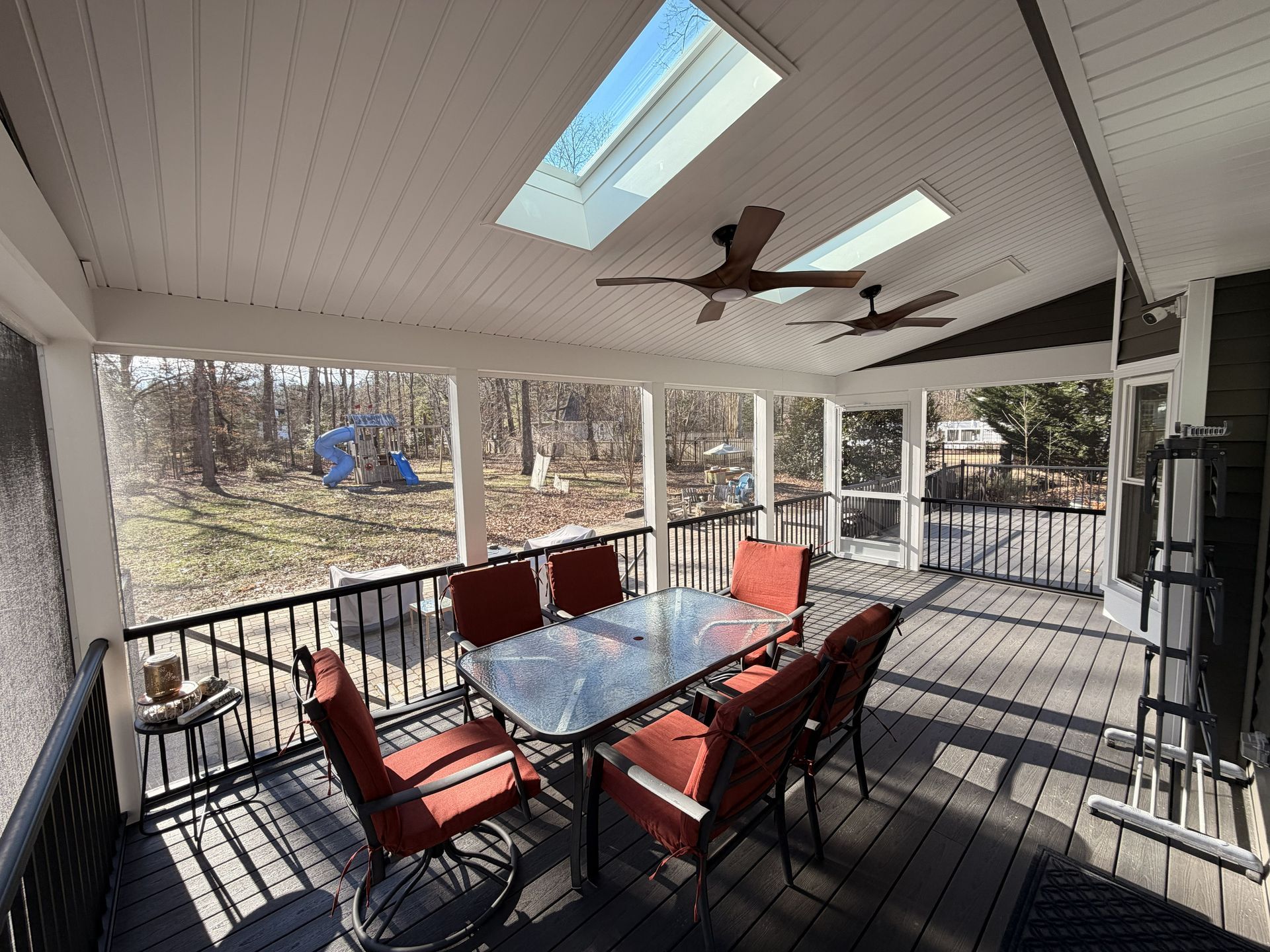 Covered porch with glass patio table, red chairs, ceiling fans, and wooded yard view.