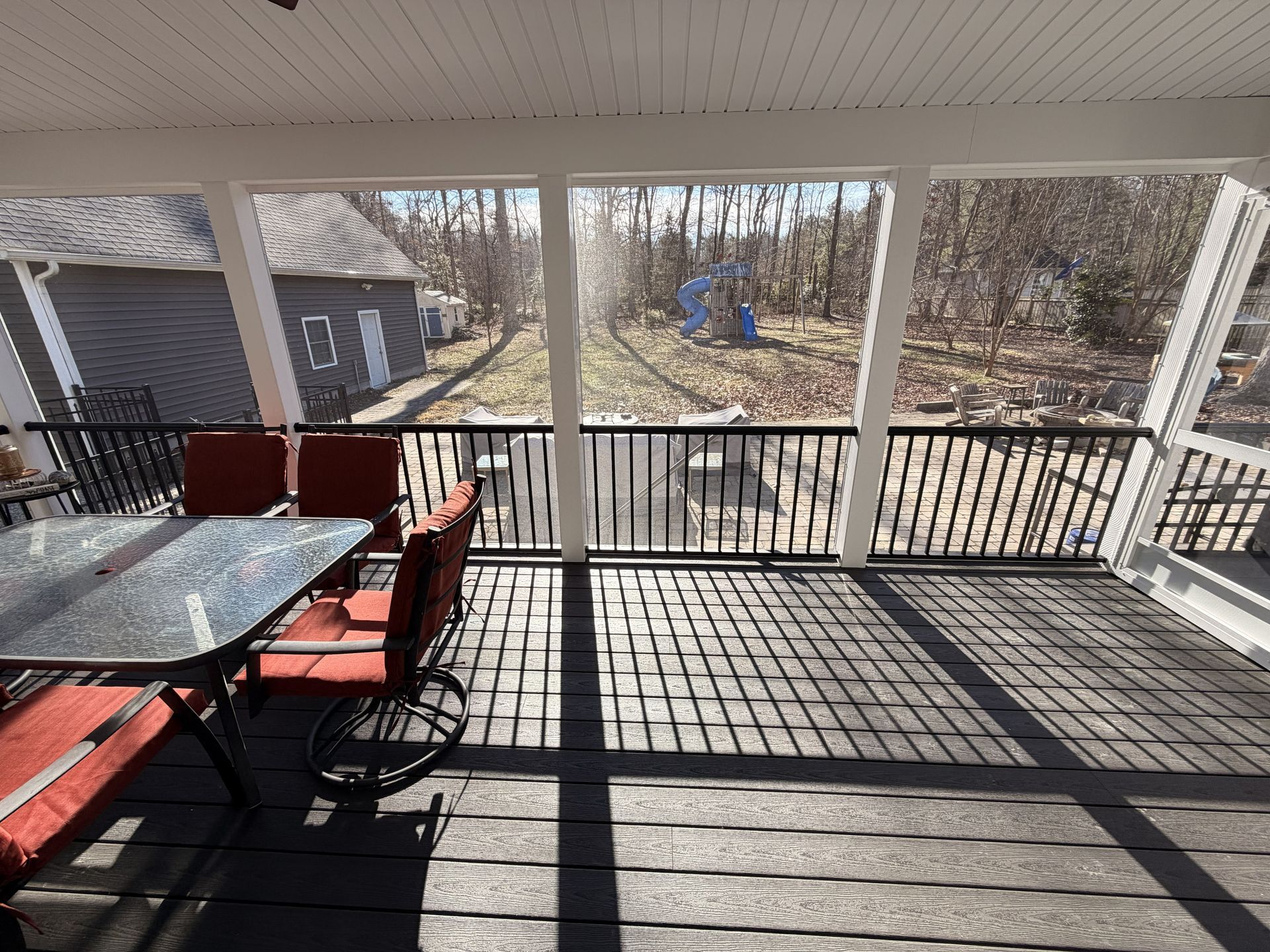 Covered deck with black railing, red chairs, and glass table overlooking a wooded yard.