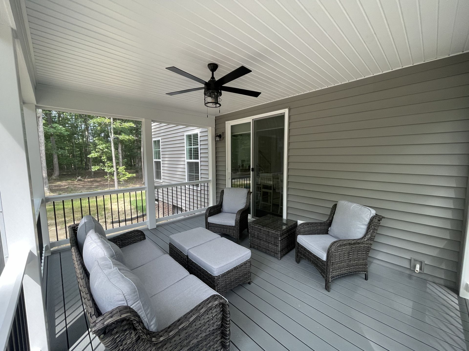Covered patio with gray outdoor furniture, ceiling fan, and sliding glass door overlooking a wooded yard
