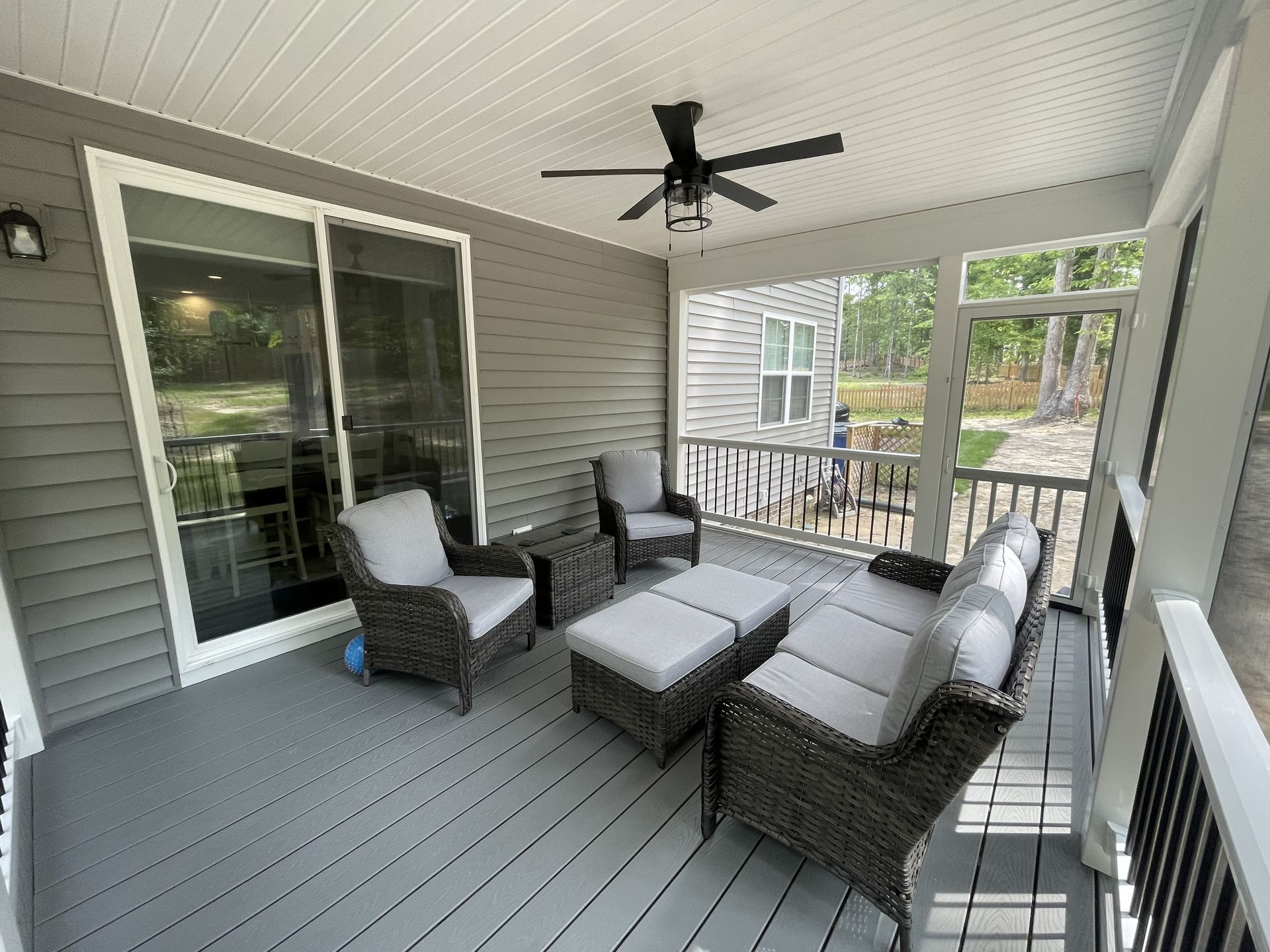 Covered porch with gray wicker seating, white cushions, and a ceiling fan overlooking a deck and trees