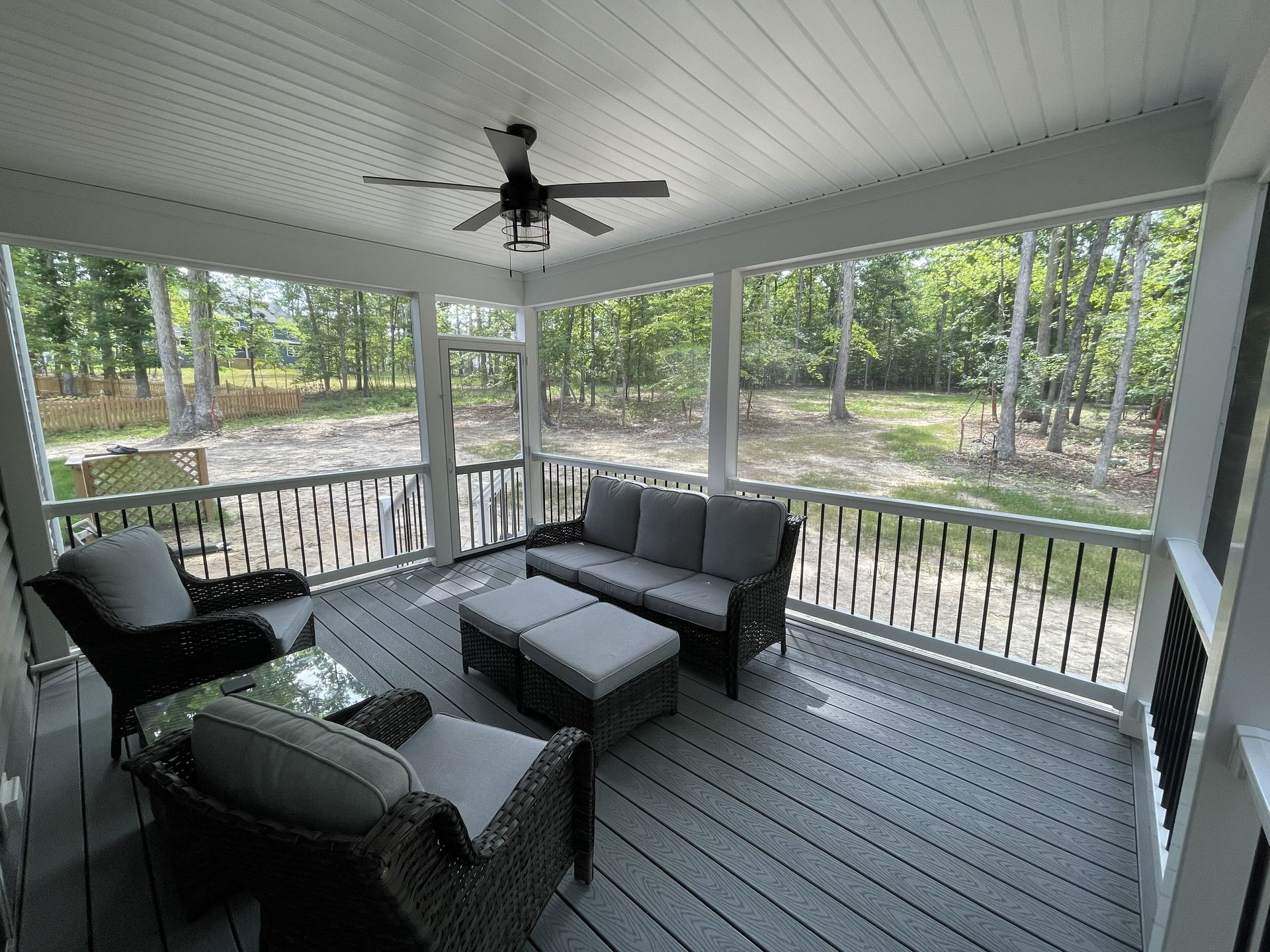 Covered porch with gray wicker seating, coffee table, and ceiling fan overlooking a wooded yard