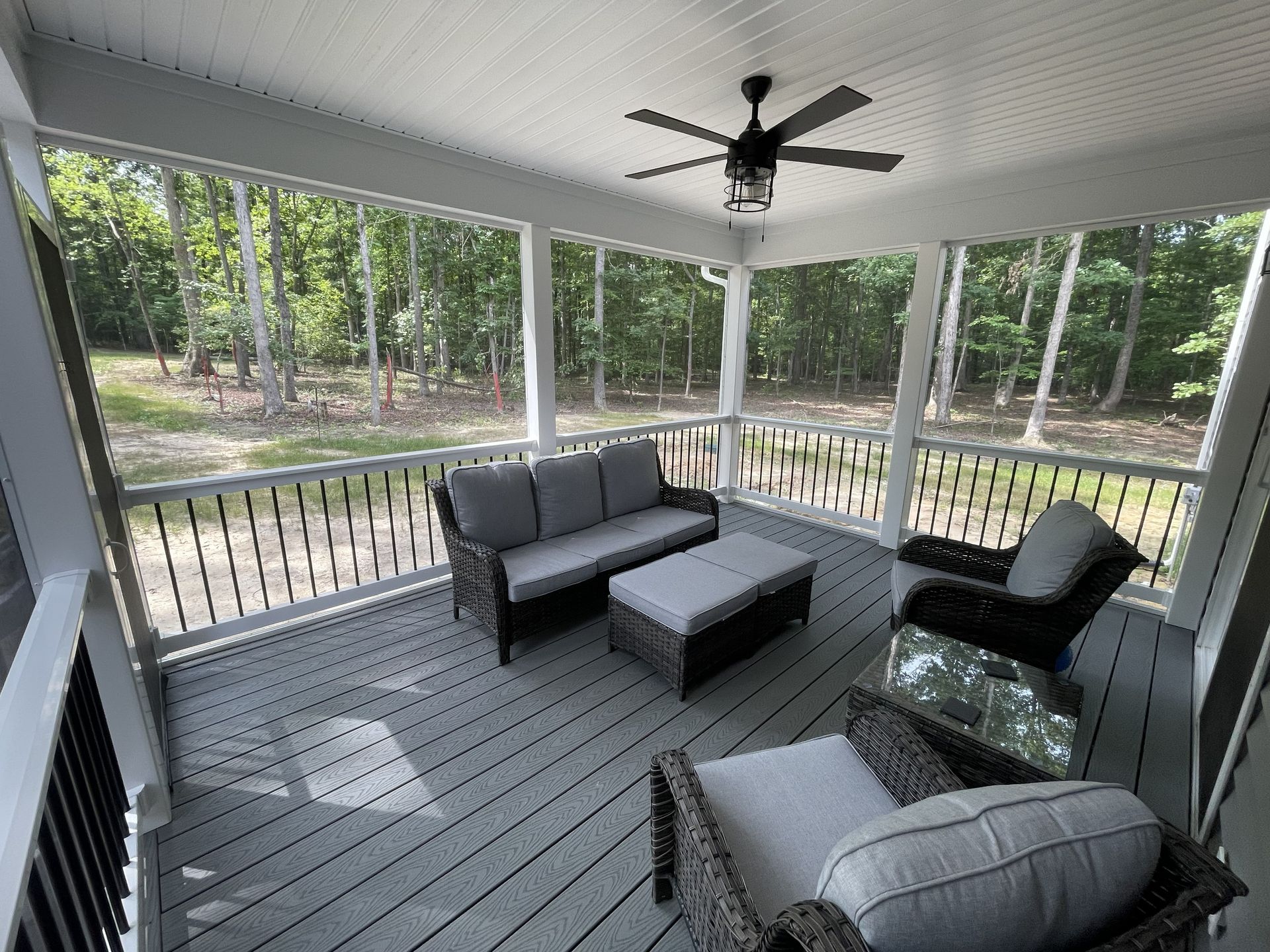 Covered porch with gray outdoor seating, ceiling fan, and wooded yard view.
