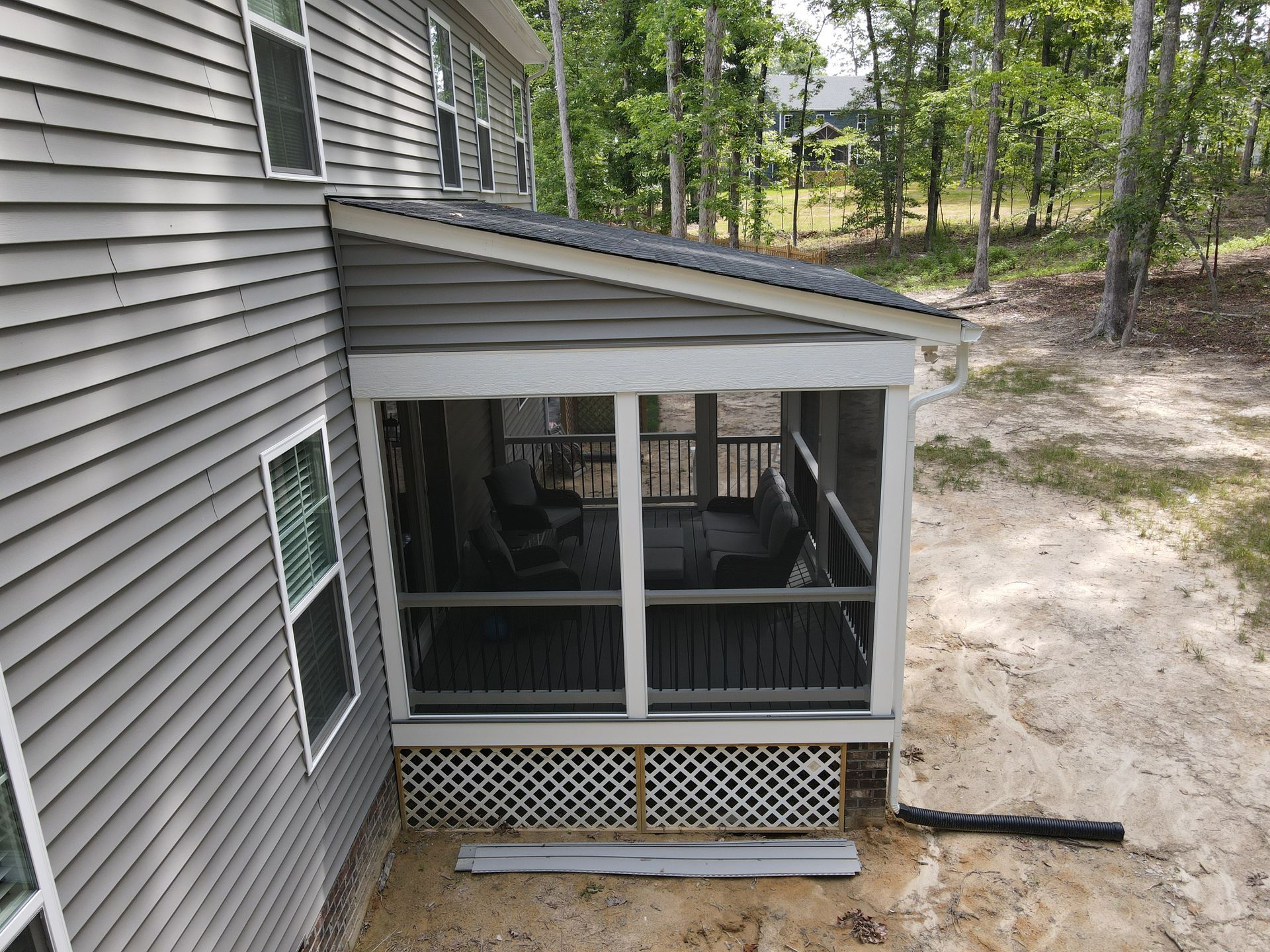 Screened porch attached to a gray house, with a sloped roof and wooded yard beyond.