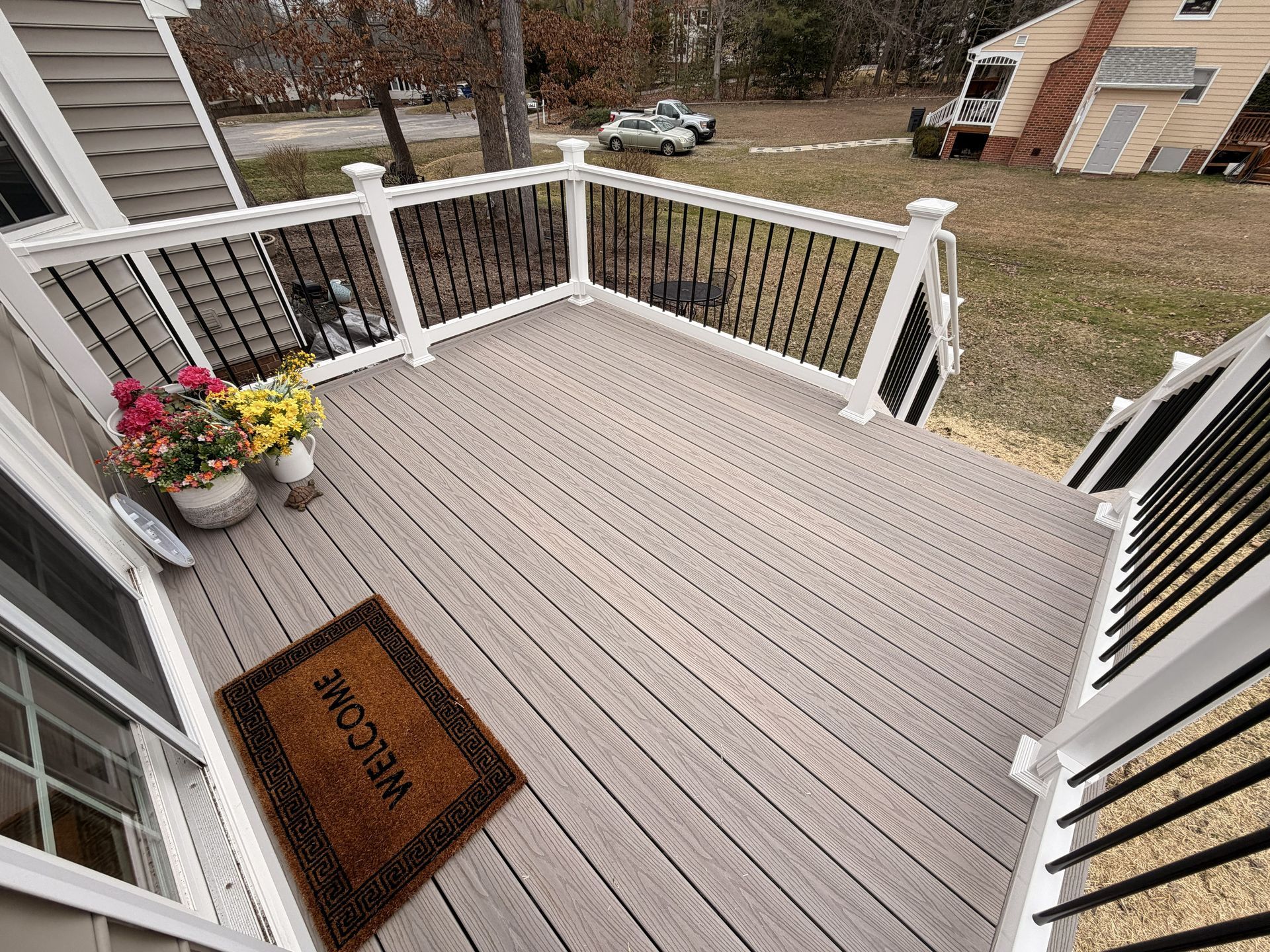 Small front porch with gray decking, white railing, flower basket, and a welcome mat facing the yard.