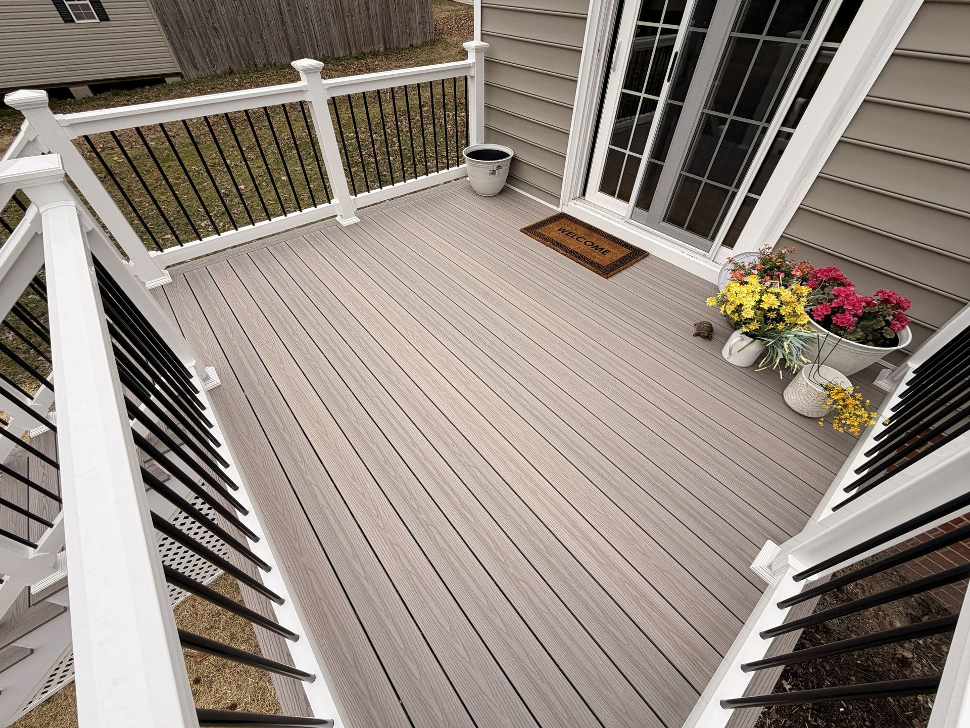 Front porch with gray decking, white railings, front door mat, and potted flowers by the entrance.
