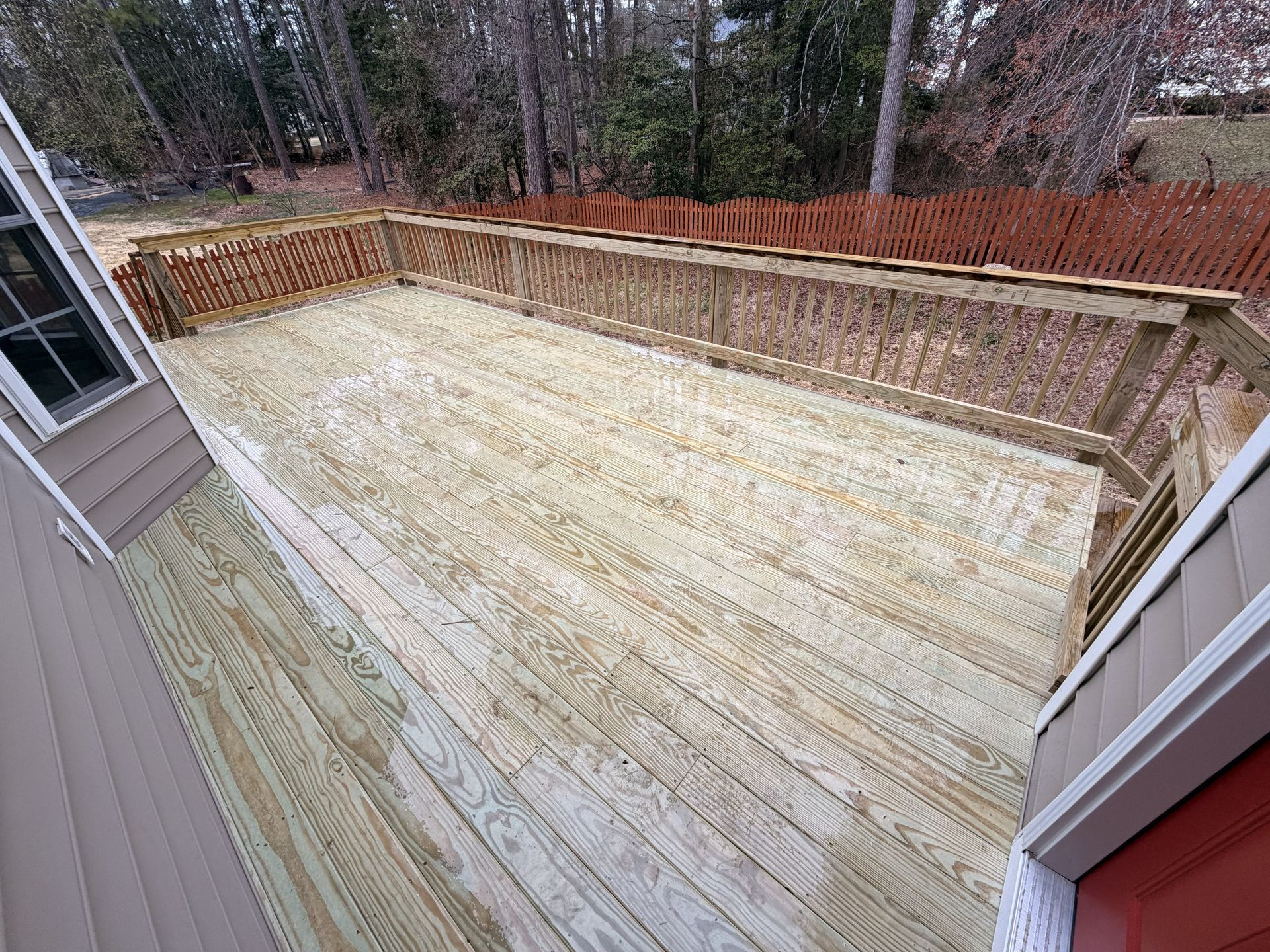 Empty wooden deck under construction, seen from a house doorway, with brick retaining wall and trees beyond