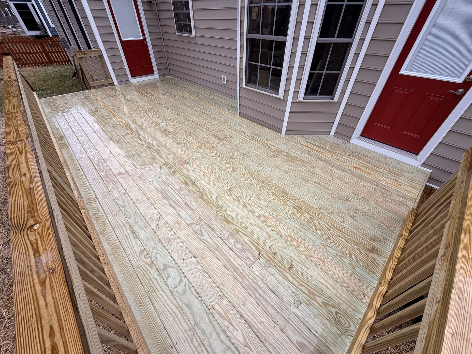 Wooden deck under construction with tan siding, red doors, and a rail along one side