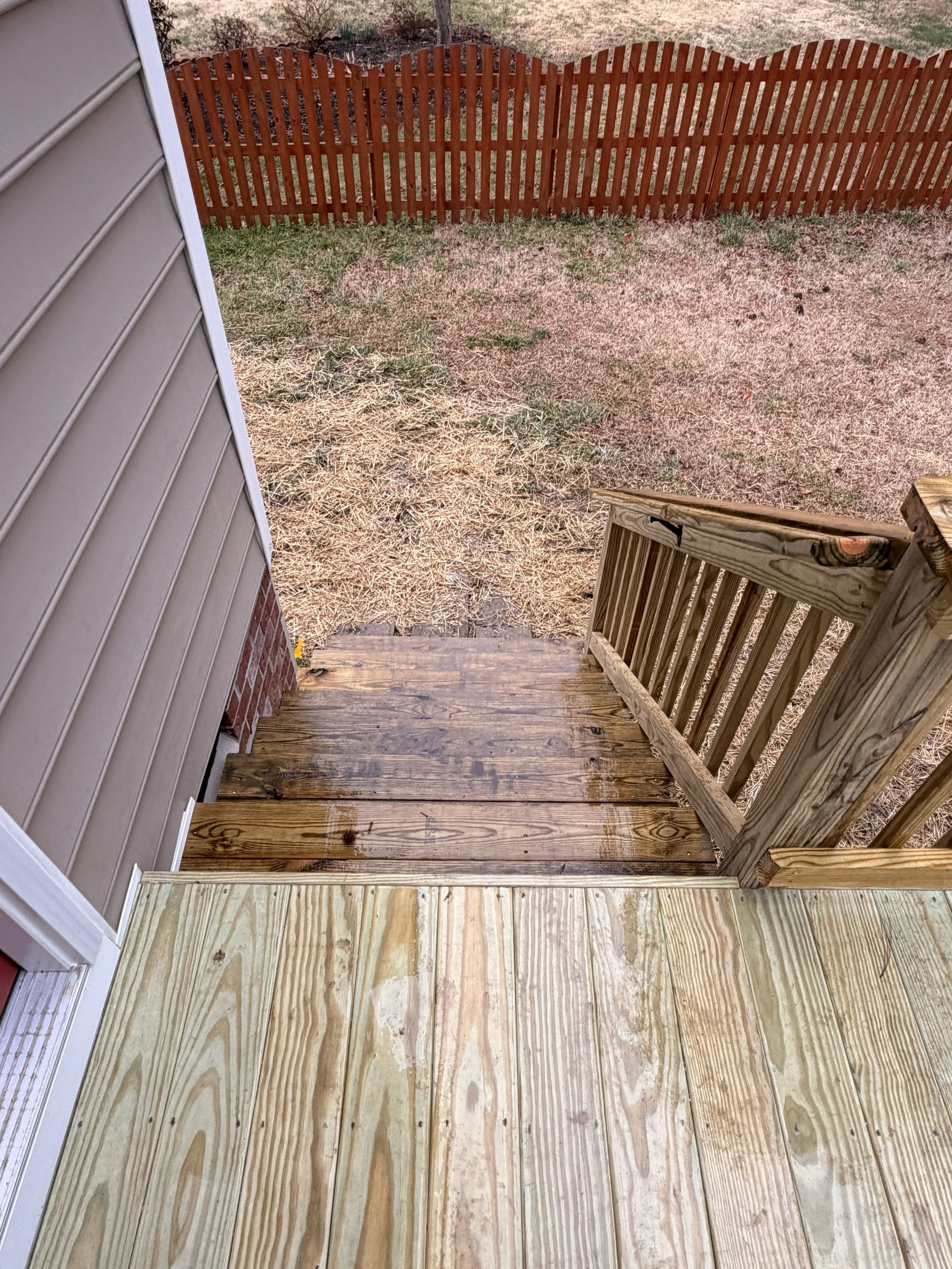 Wooden deck stairs descending to a patchy backyard with a red fence and house siding on the left