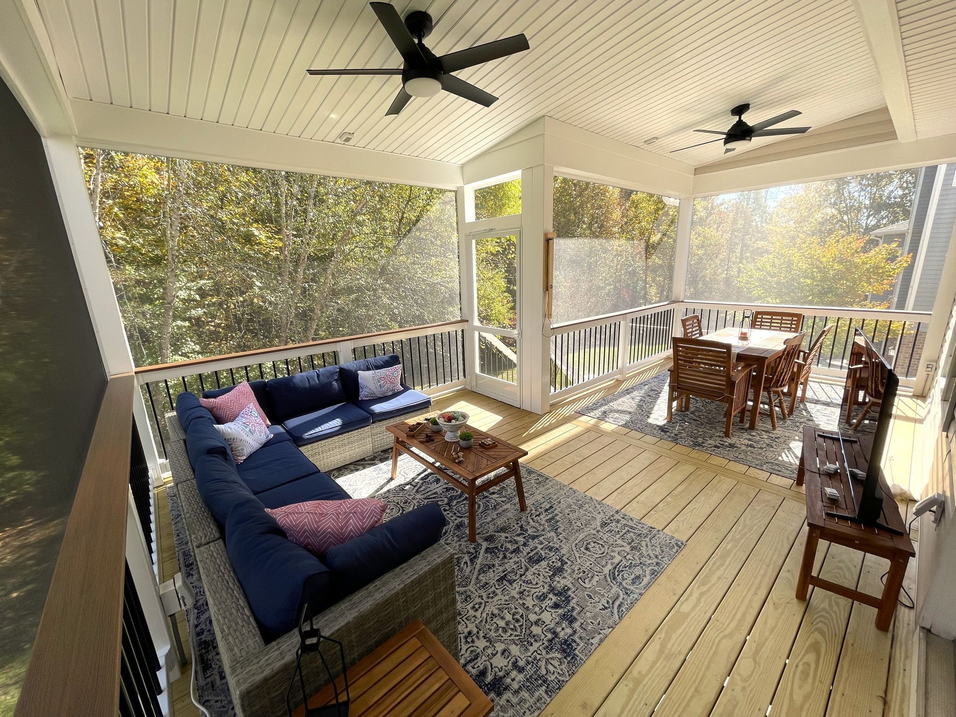 Covered porch with sectional sofa, dining table, and ceiling fans overlooking trees
