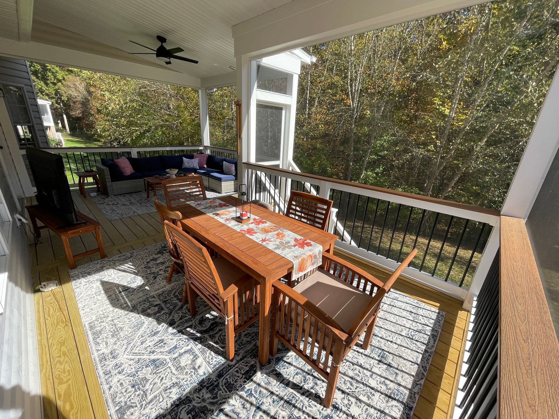 Covered porch dining area with wooden table, chairs, rug, and view of trees outside