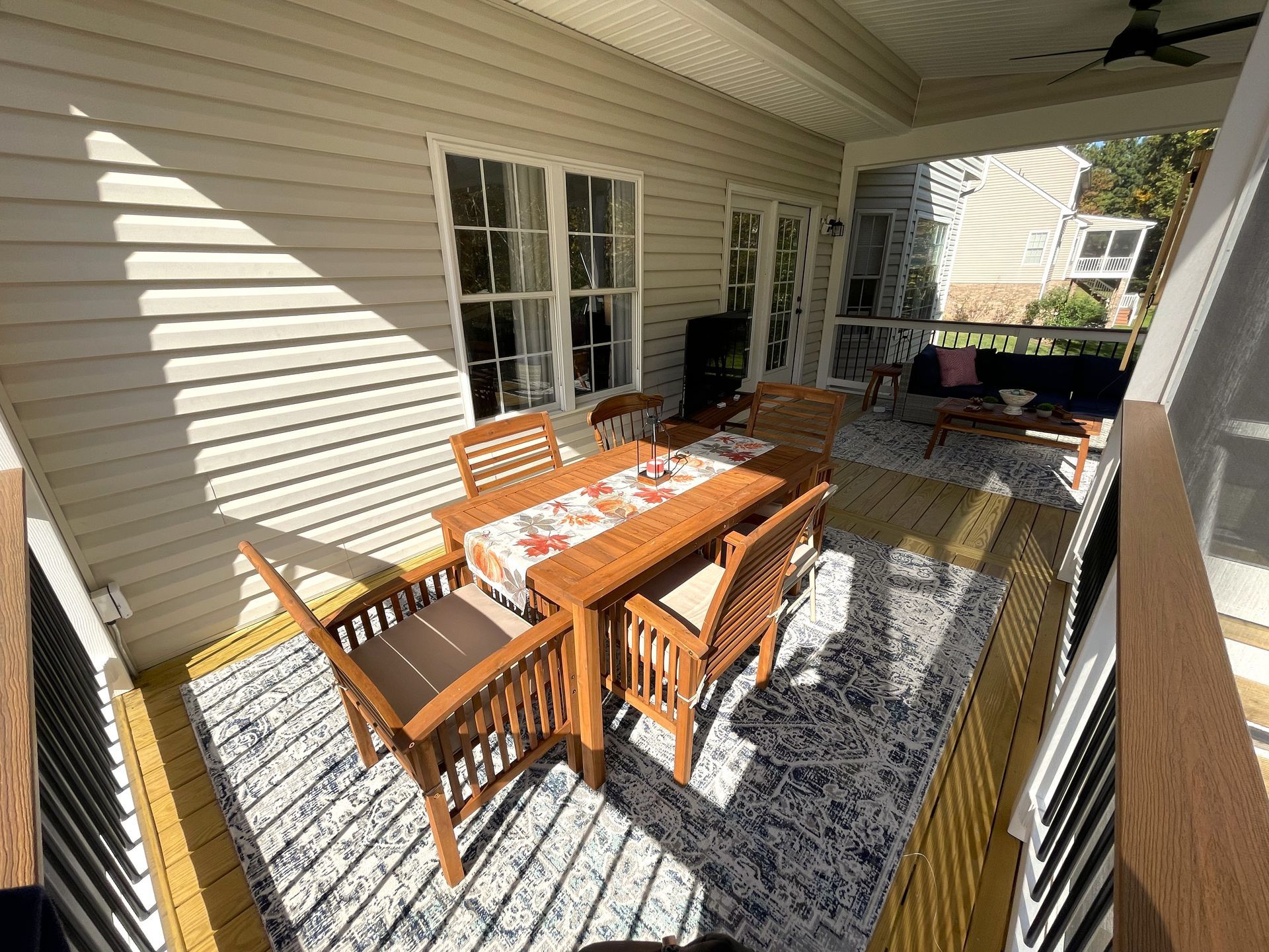 Covered patio with wooden dining table and chairs, patterned rug, and cushioned seating in bright sunlight