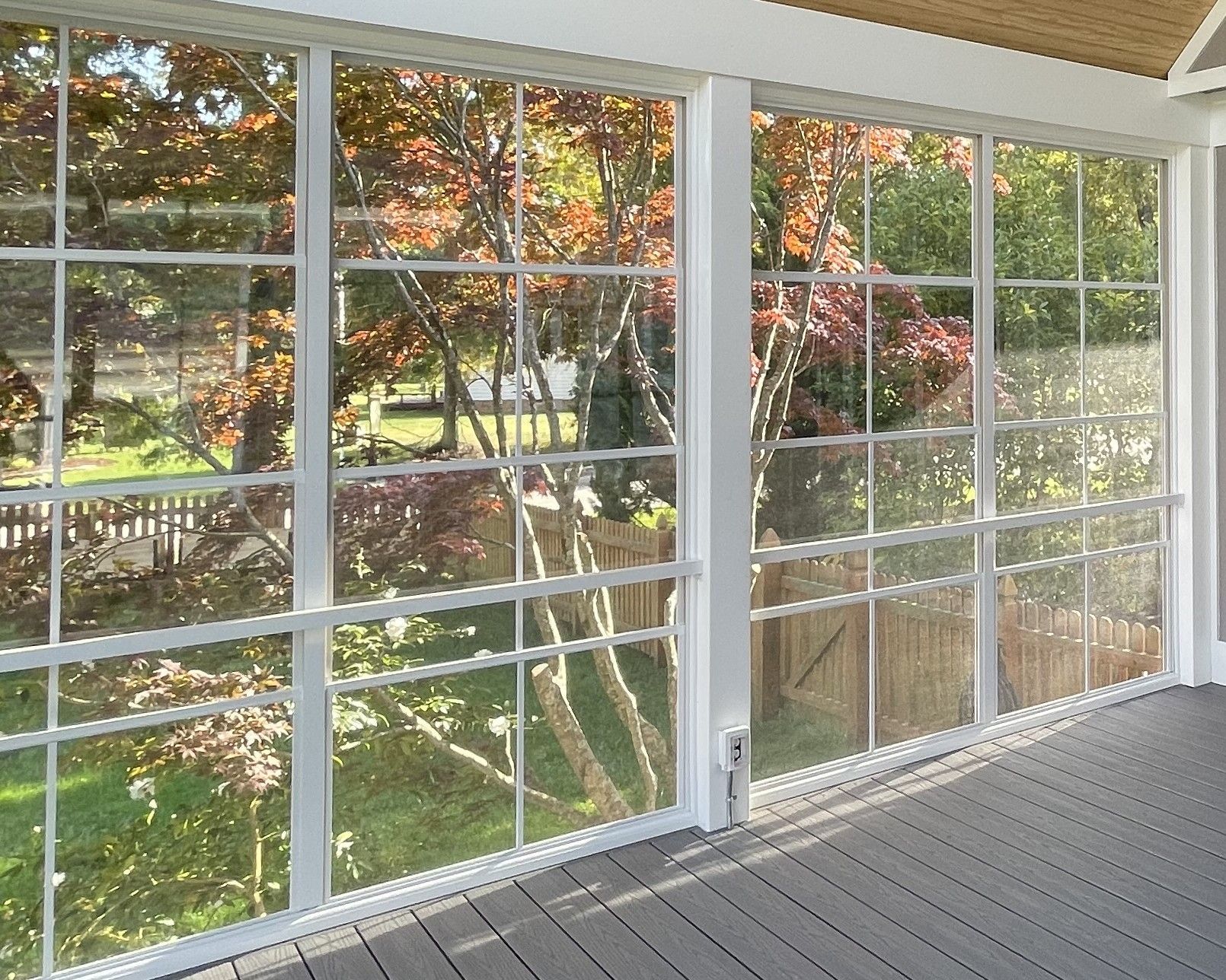 Sunroom with floor-to-ceiling windows, showing a leafy backyard and wooden deck flooring.