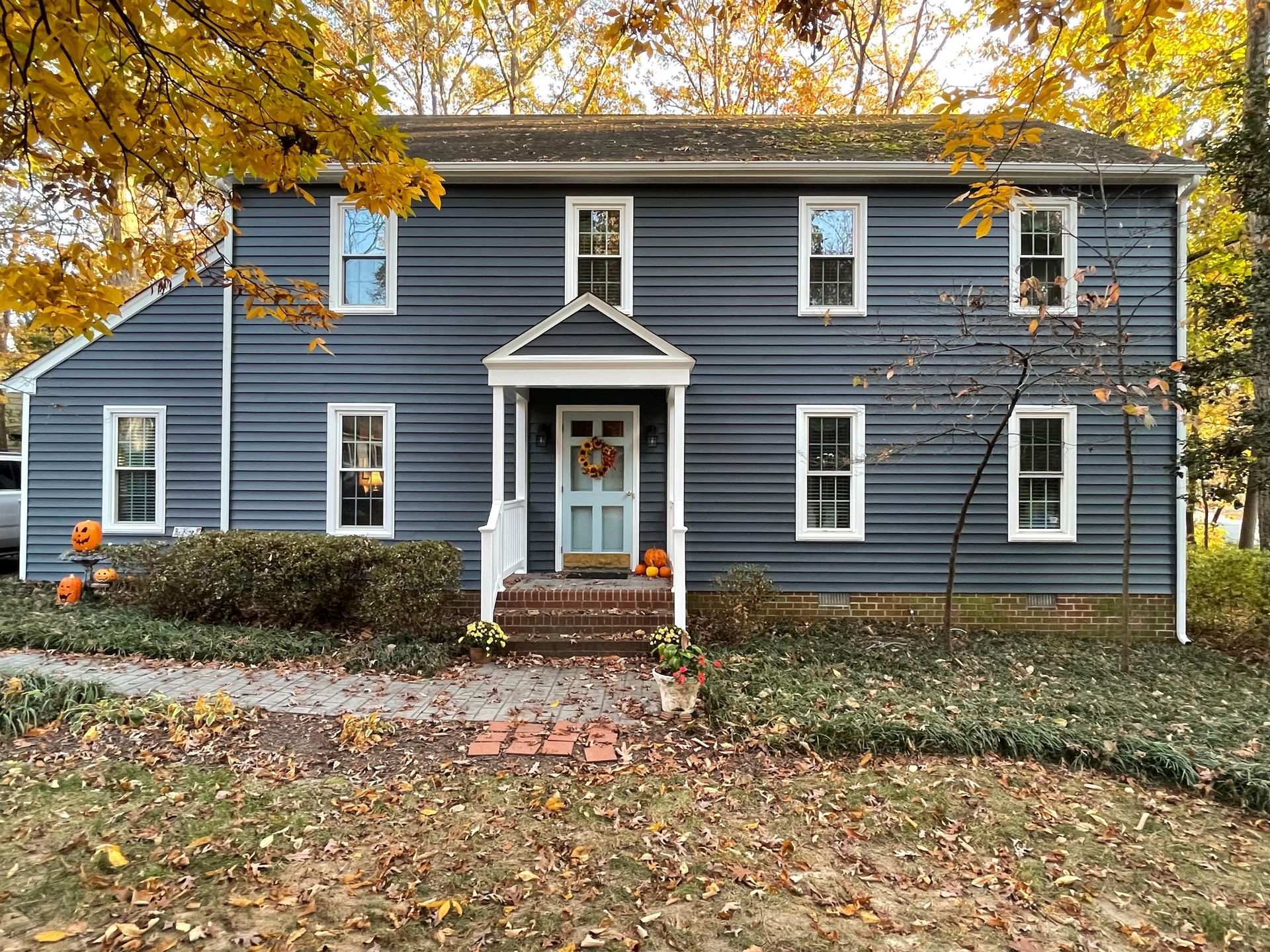 Blue two-story house with white trim and a small porch, framed by autumn trees and fallen leaves.