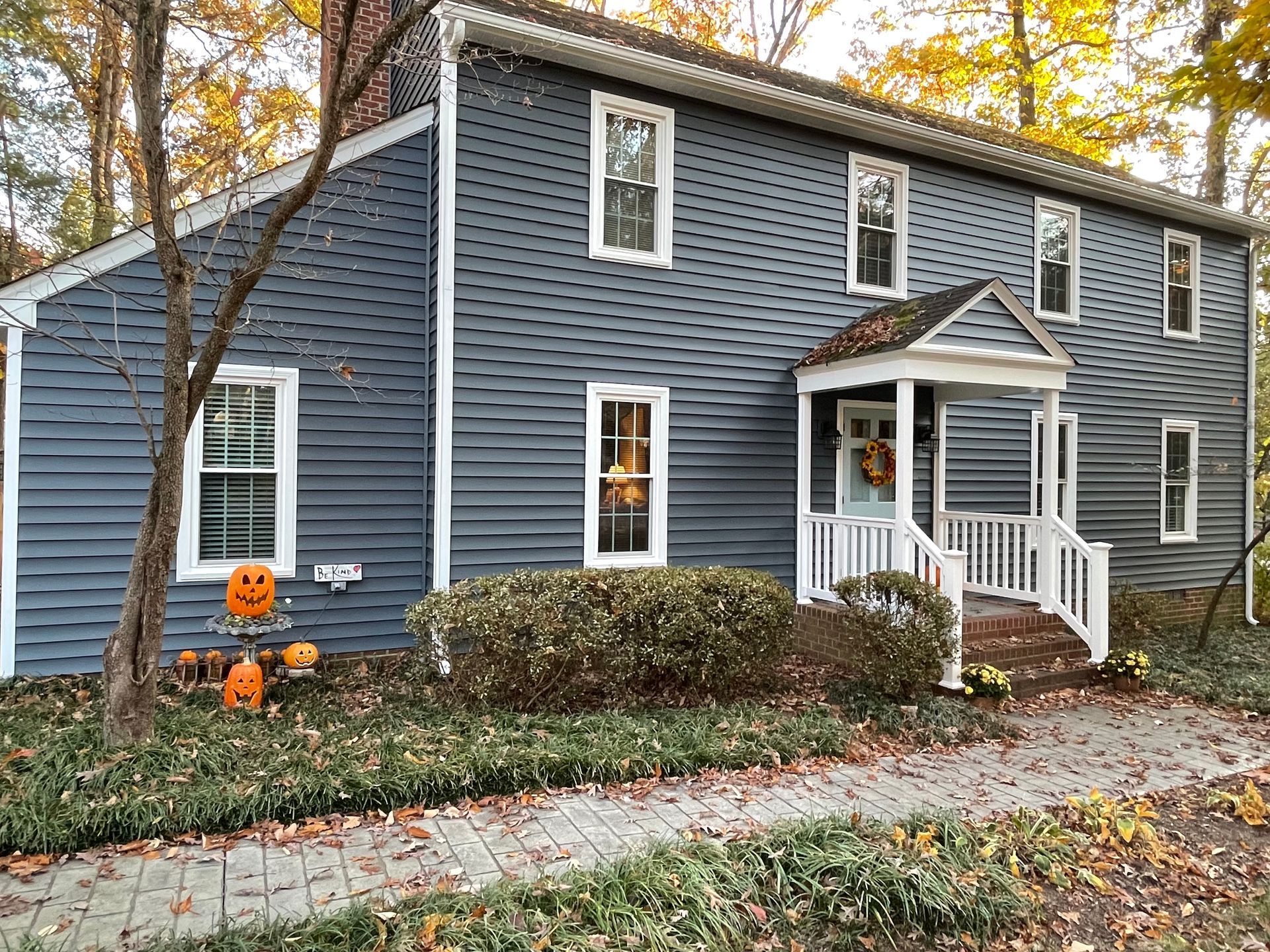 Blue two-story house with white trim, autumn trees, and Halloween pumpkins by the front steps