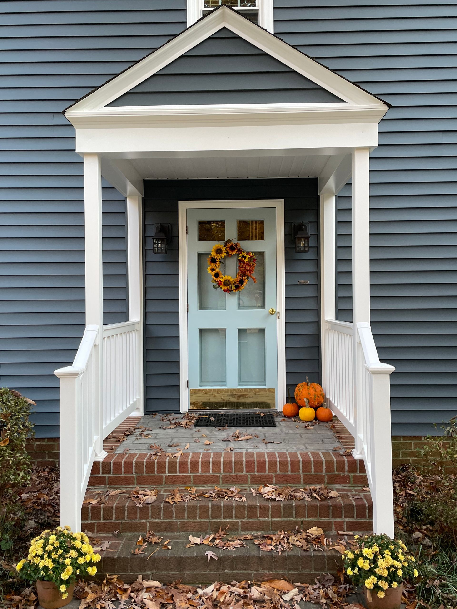 Front porch with light blue door, white trim, fall wreath, pumpkins, and potted mums on brick steps