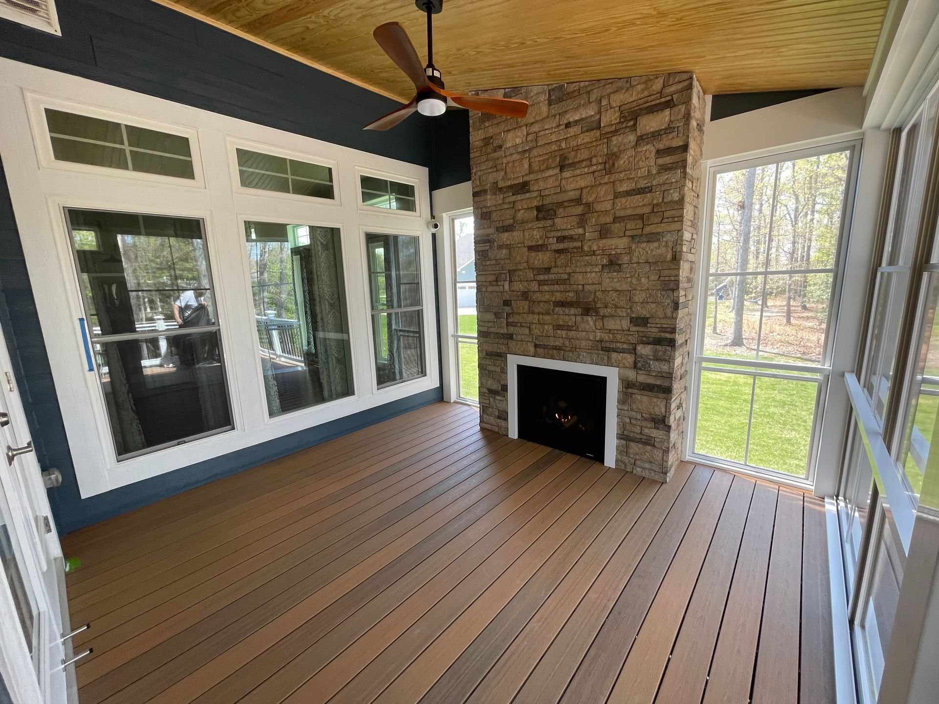 Screened porch with wood floor, stone fireplace, ceiling fan, and large windows overlooking a yard