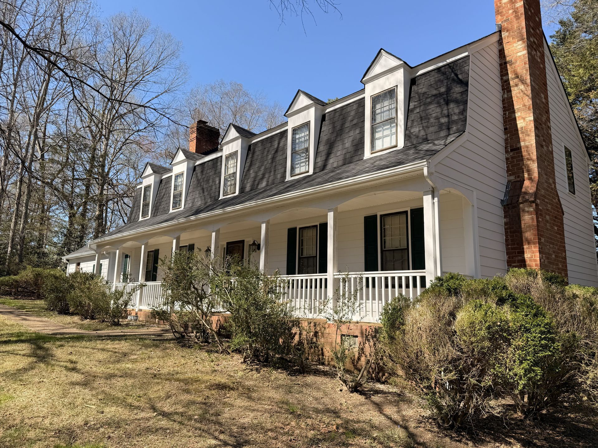 White farmhouse with dormer windows, front porch, and brick chimney on a sunny wooded lot.