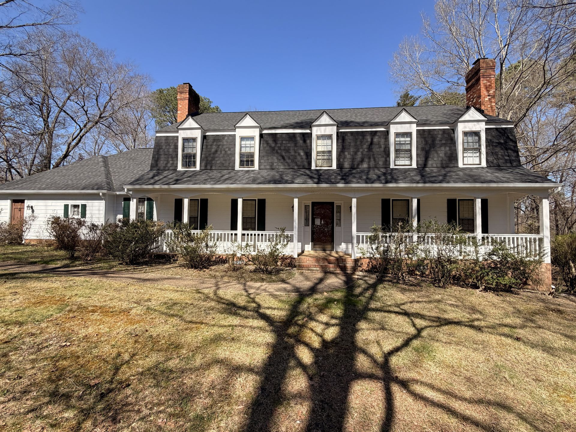 Large white farmhouse with a wraparound porch, dark roof, and bare trees casting shadows on the lawn.