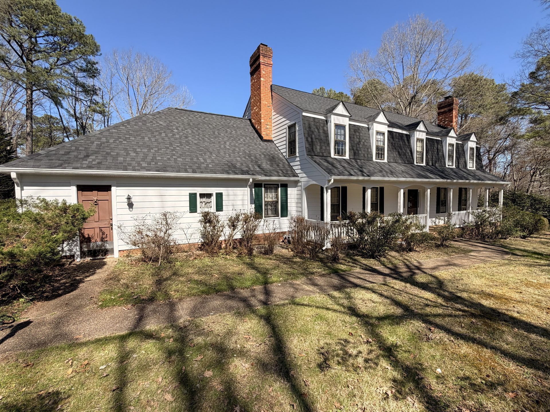 Large white house with gray roof, brick chimney, and front porch in a sunny yard with trees