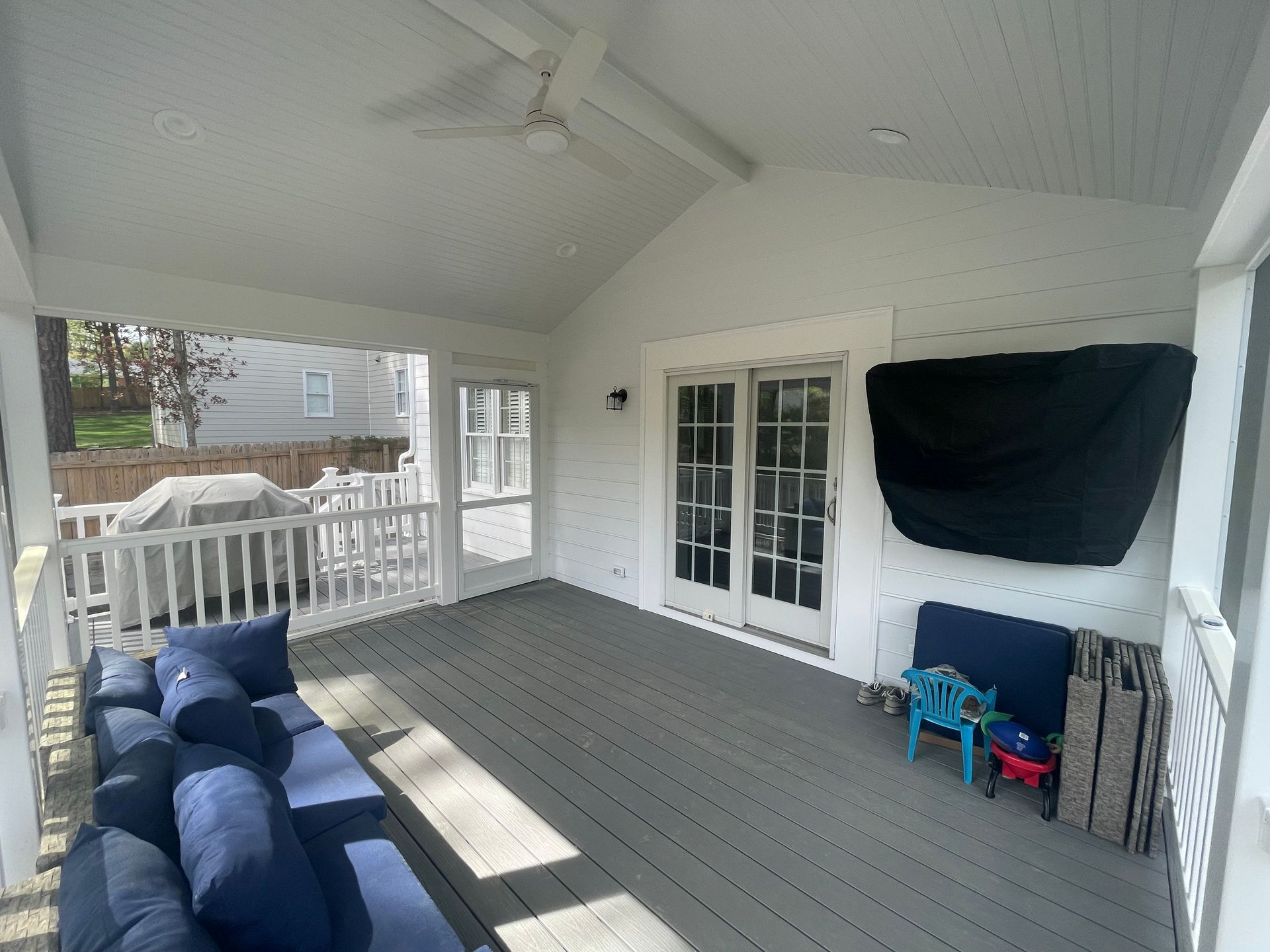 Covered porch with blue cushioned chairs, white railing, French doors, and a covered TV on the wall.
