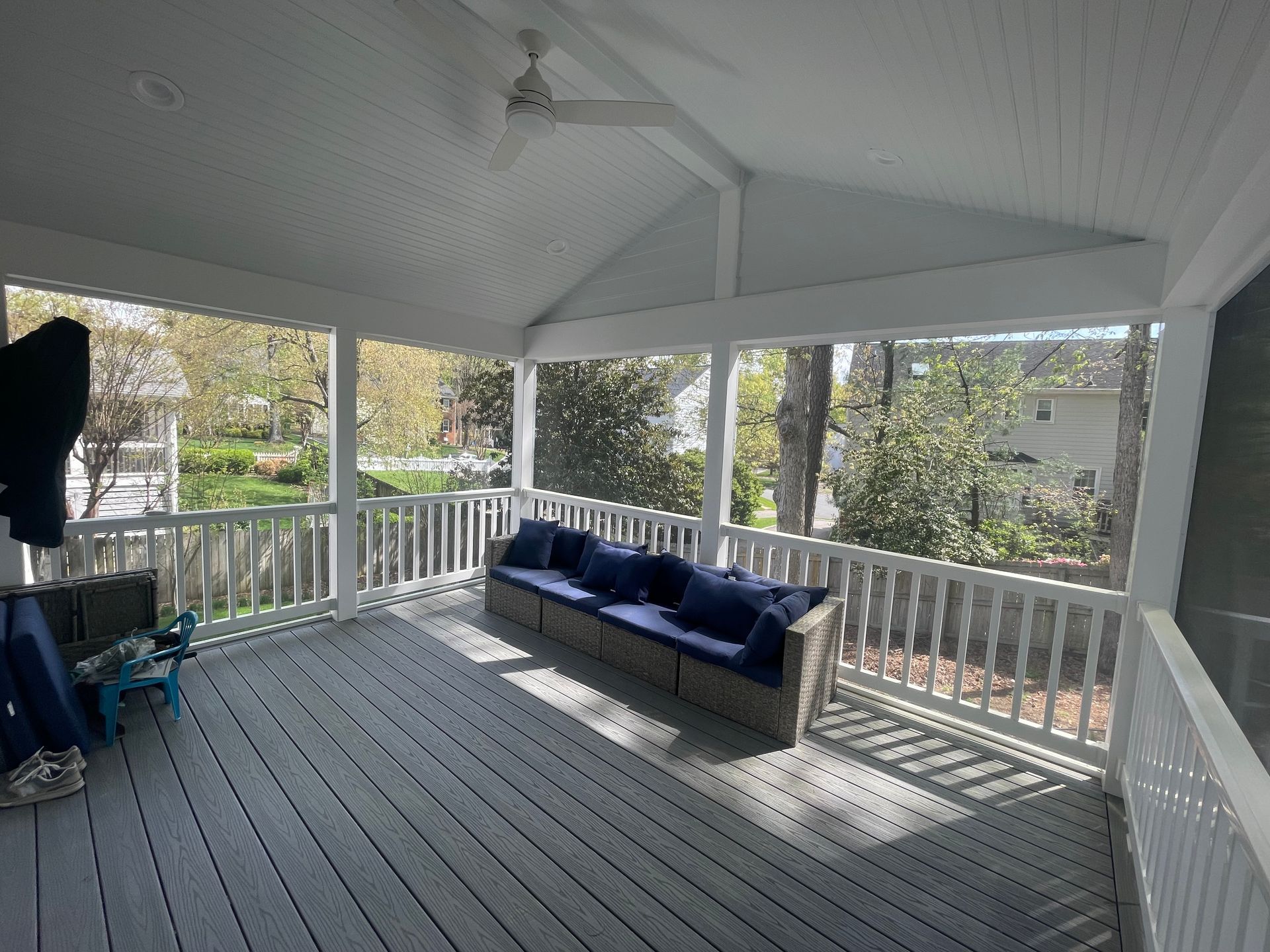 Covered porch with gray floorboards, white railings, and a blue sofa in sunlight.