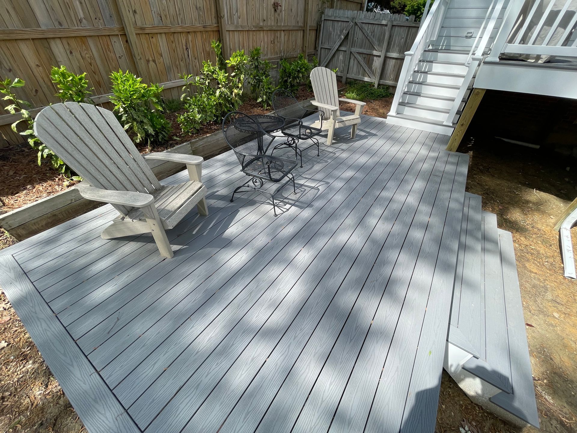 Gray deck with Adirondack chairs and a small table in a fenced backyard, beside stairs and a raised porch