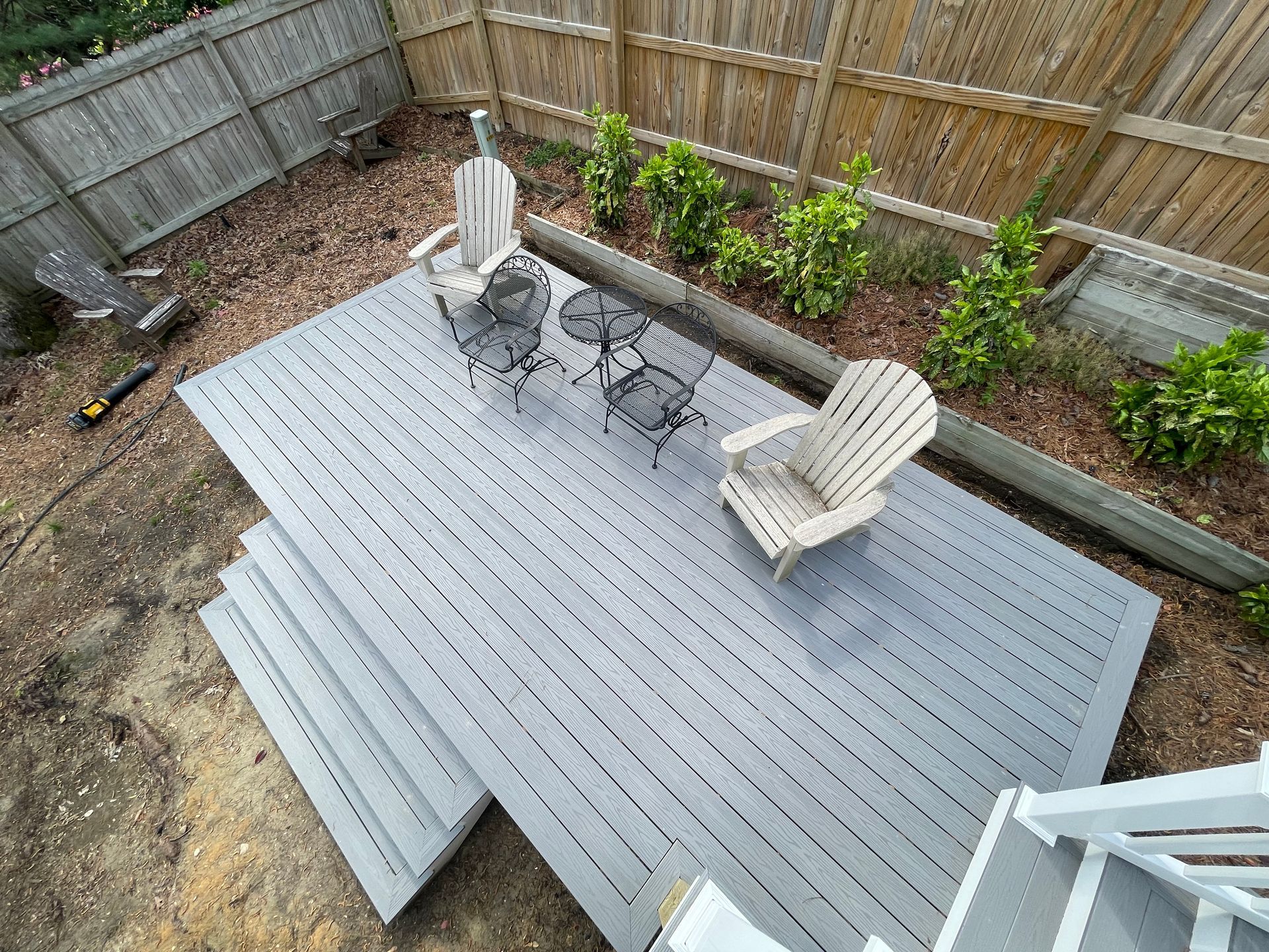Backyard deck with gray seating, two chairs and a table, bordered by a wooden fence and garden beds
