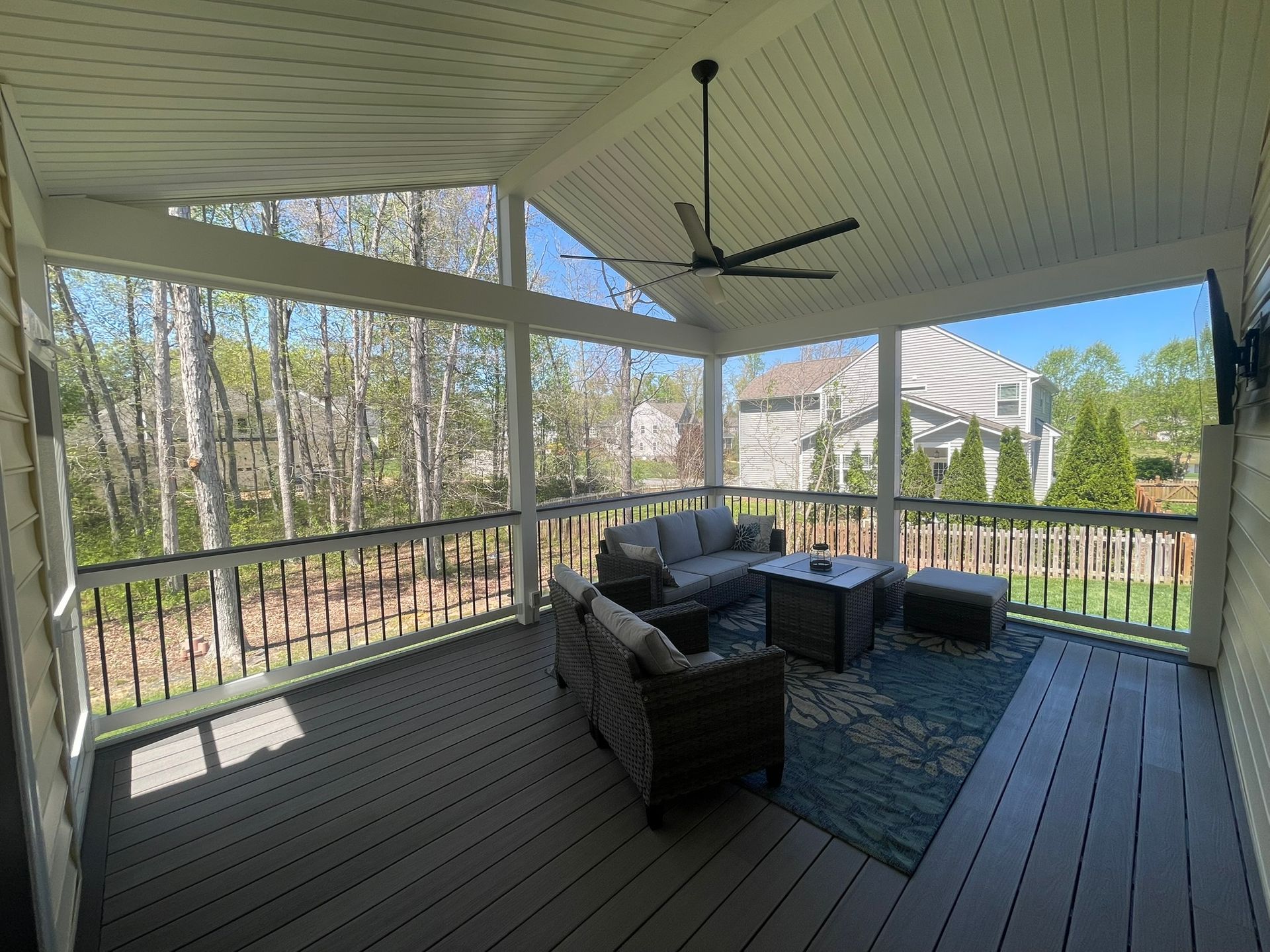 Screened porch with wicker seating, blue rug, ceiling fan, and wooded backyard view