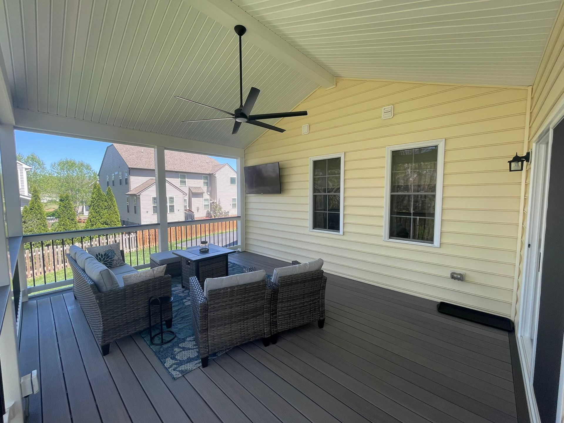 Covered porch with gray wicker seating, ceiling fan, and yellow siding overlooking a neighborhood.