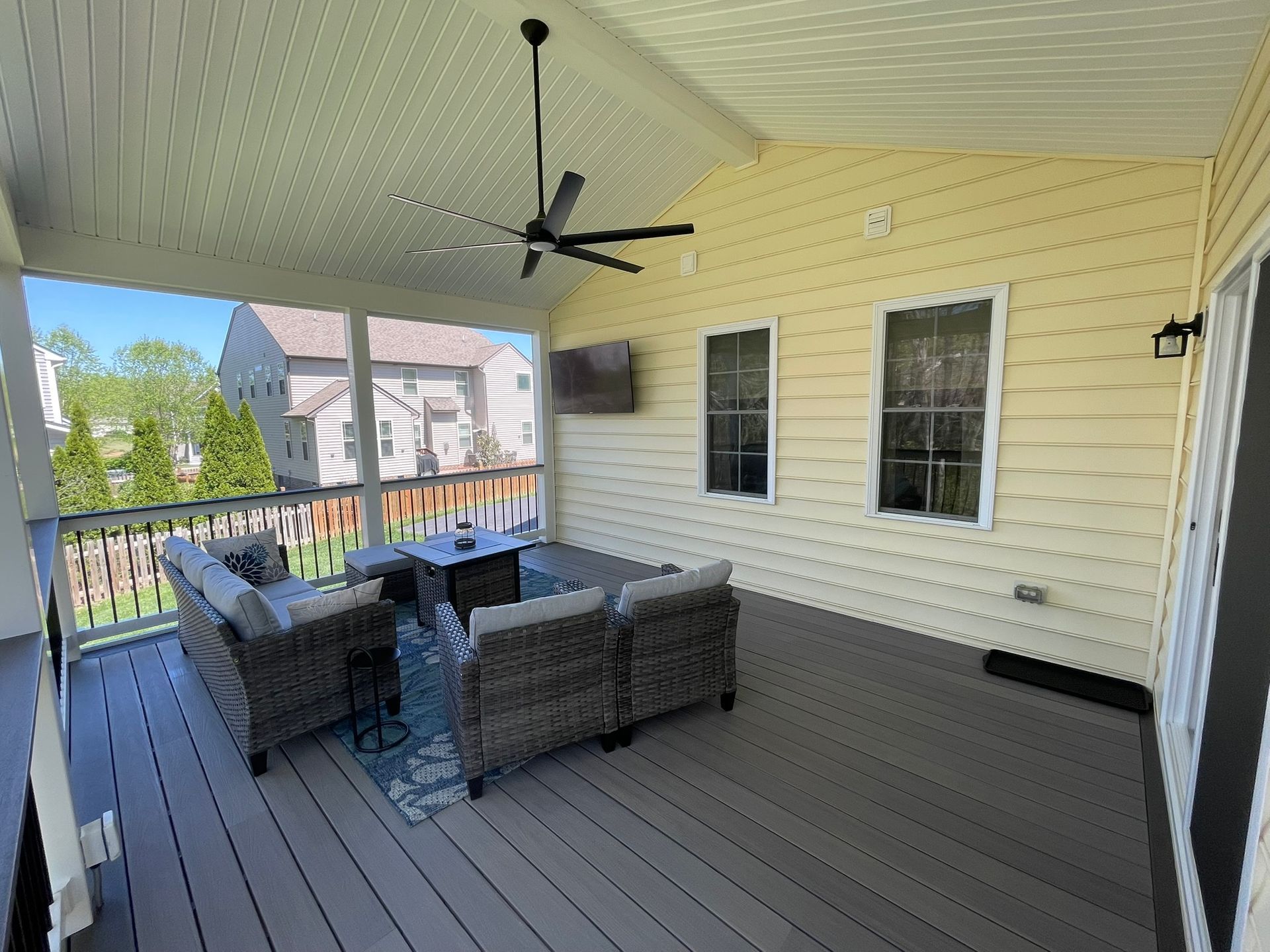 Covered porch with gray wicker seating, ceiling fan, and yellow siding overlooking neighboring homes