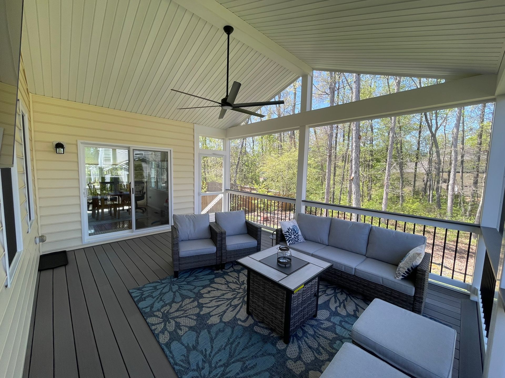 Covered porch with gray outdoor seating, ceiling fan, and wooded view through screened railing.