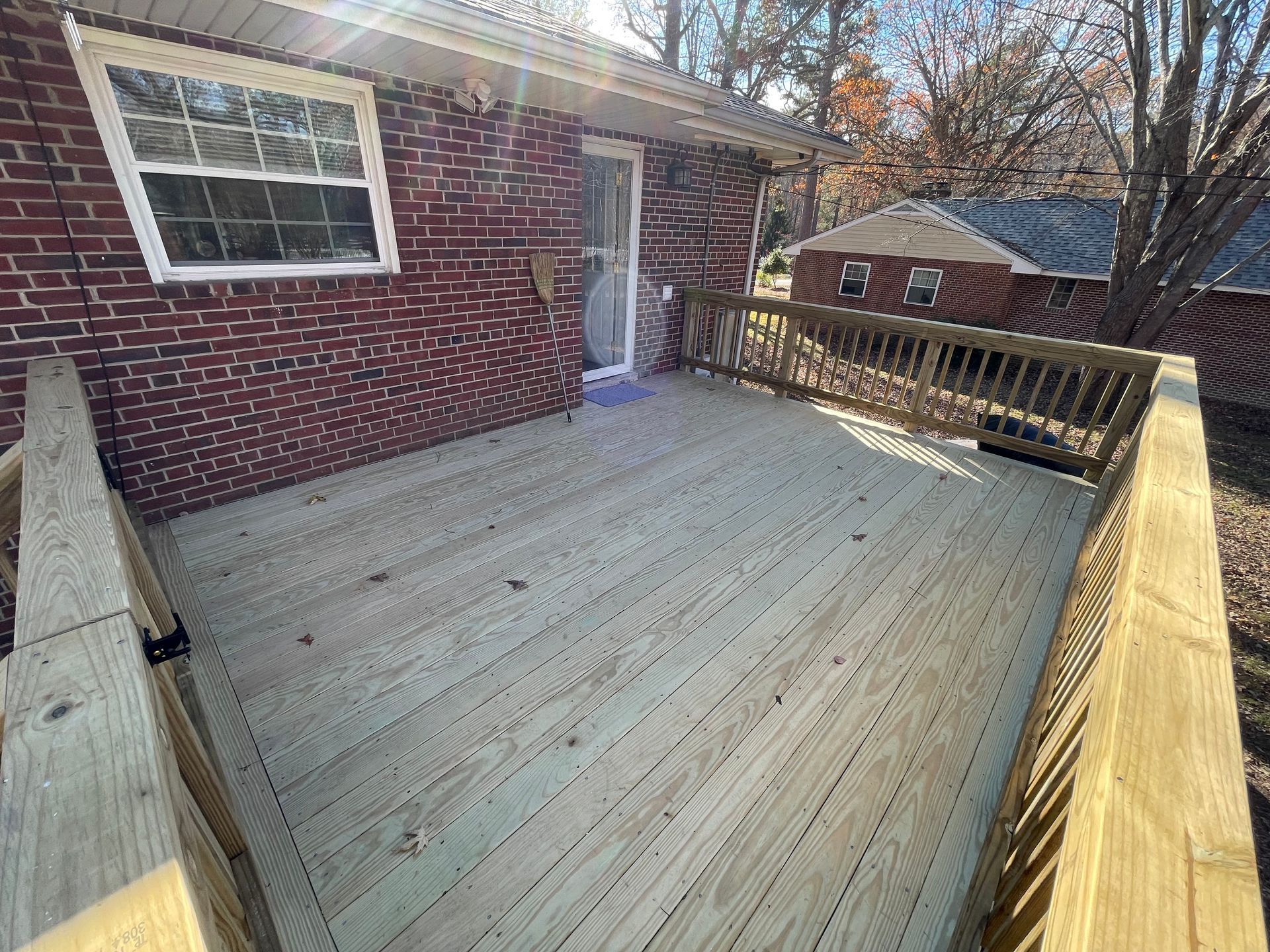 Wooden deck beside a brick house, with railing and stairs in a sunny backyard.