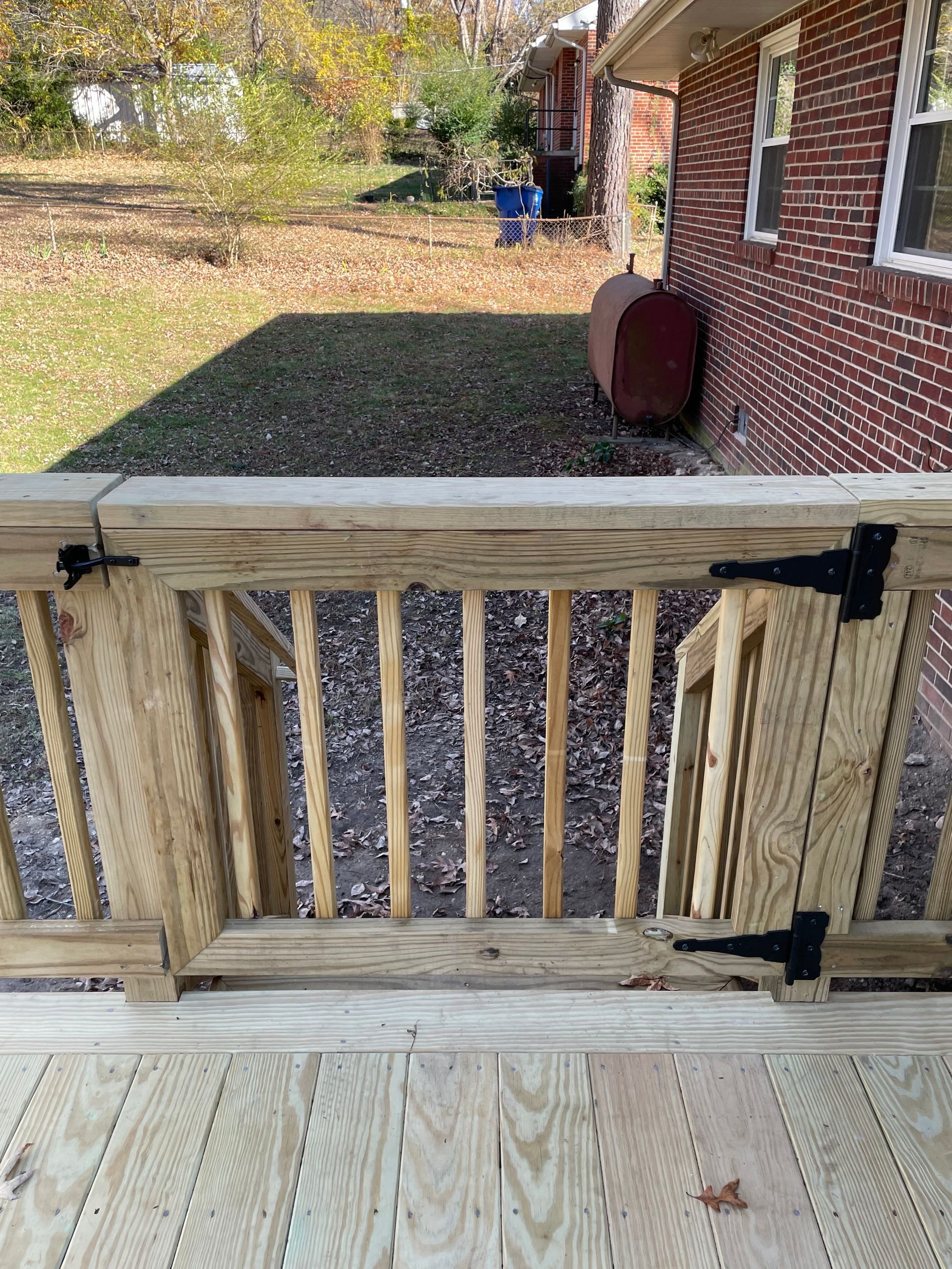 Wooden deck gate with black hinges opens onto a leaf-covered yard beside a brick house.