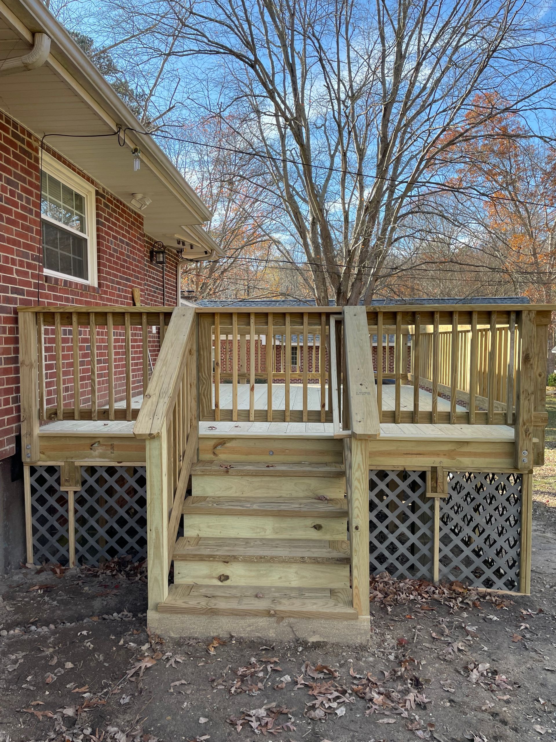 Wooden deck stairs with railings beside a brick house, framed by bare trees in a yard