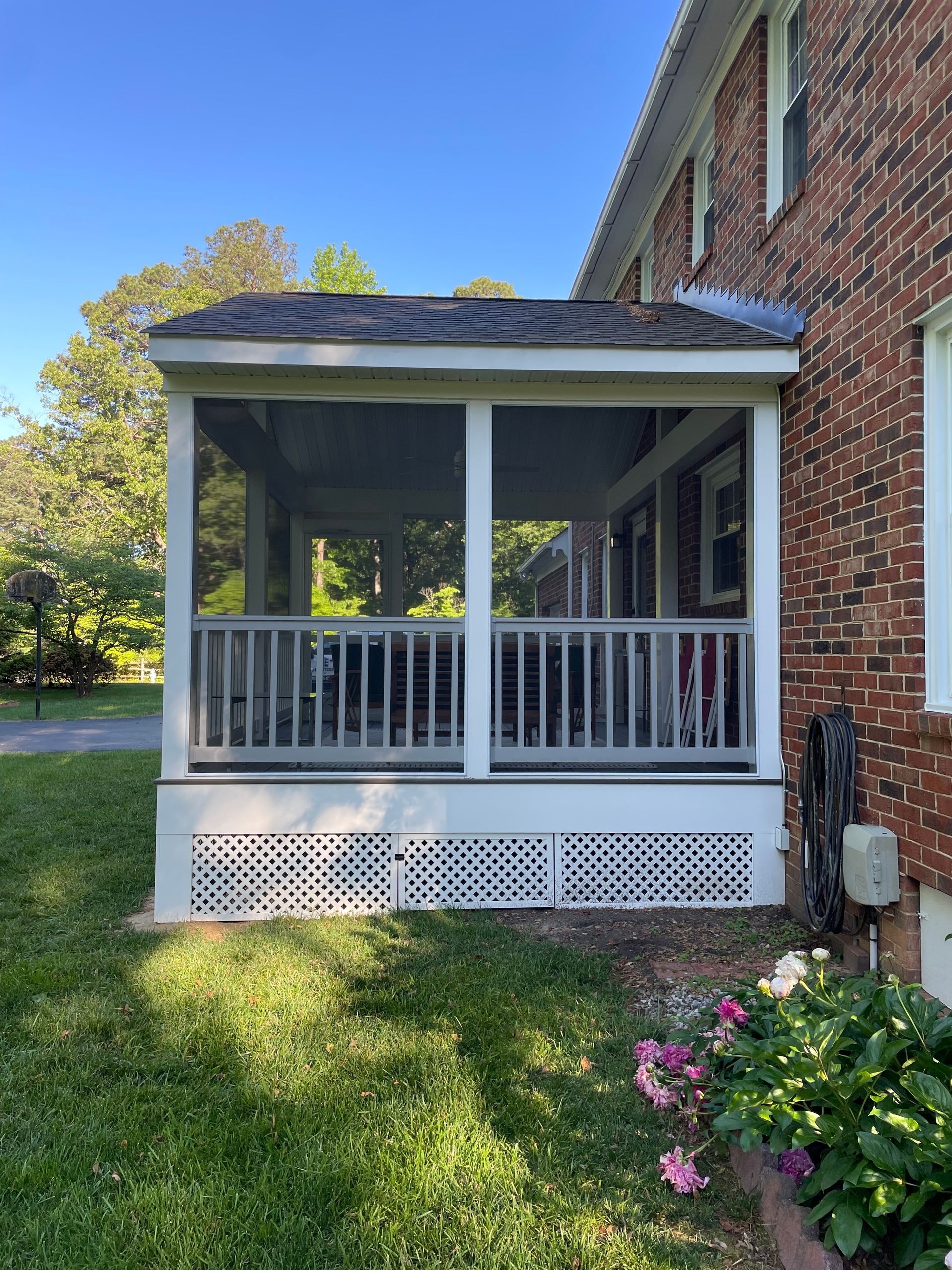 Front porch with white railings on a brick house, surrounded by green lawn and flowers.