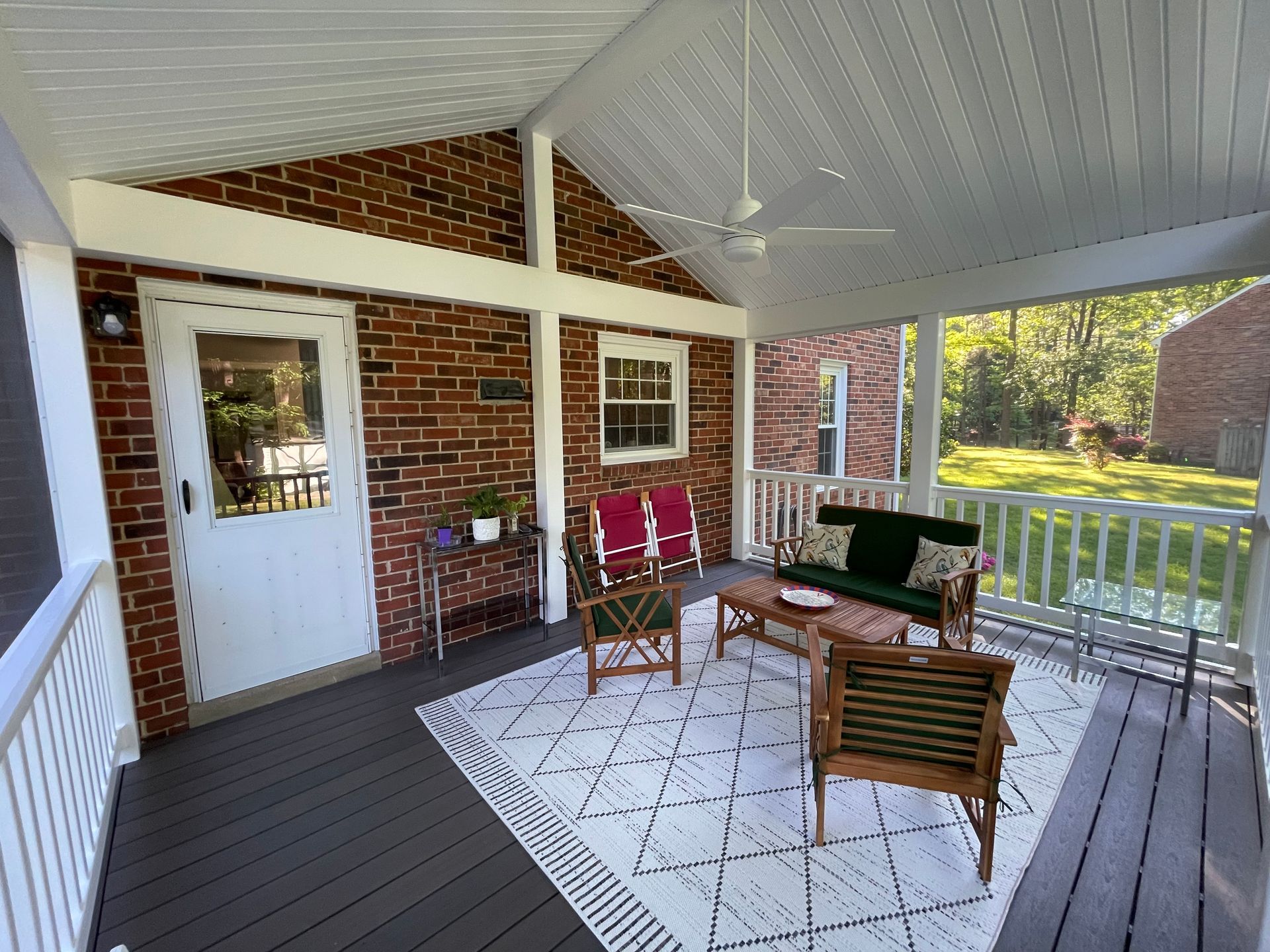 Covered porch with brick wall, white door, wicker chairs, rug, and railing overlooking a green yard