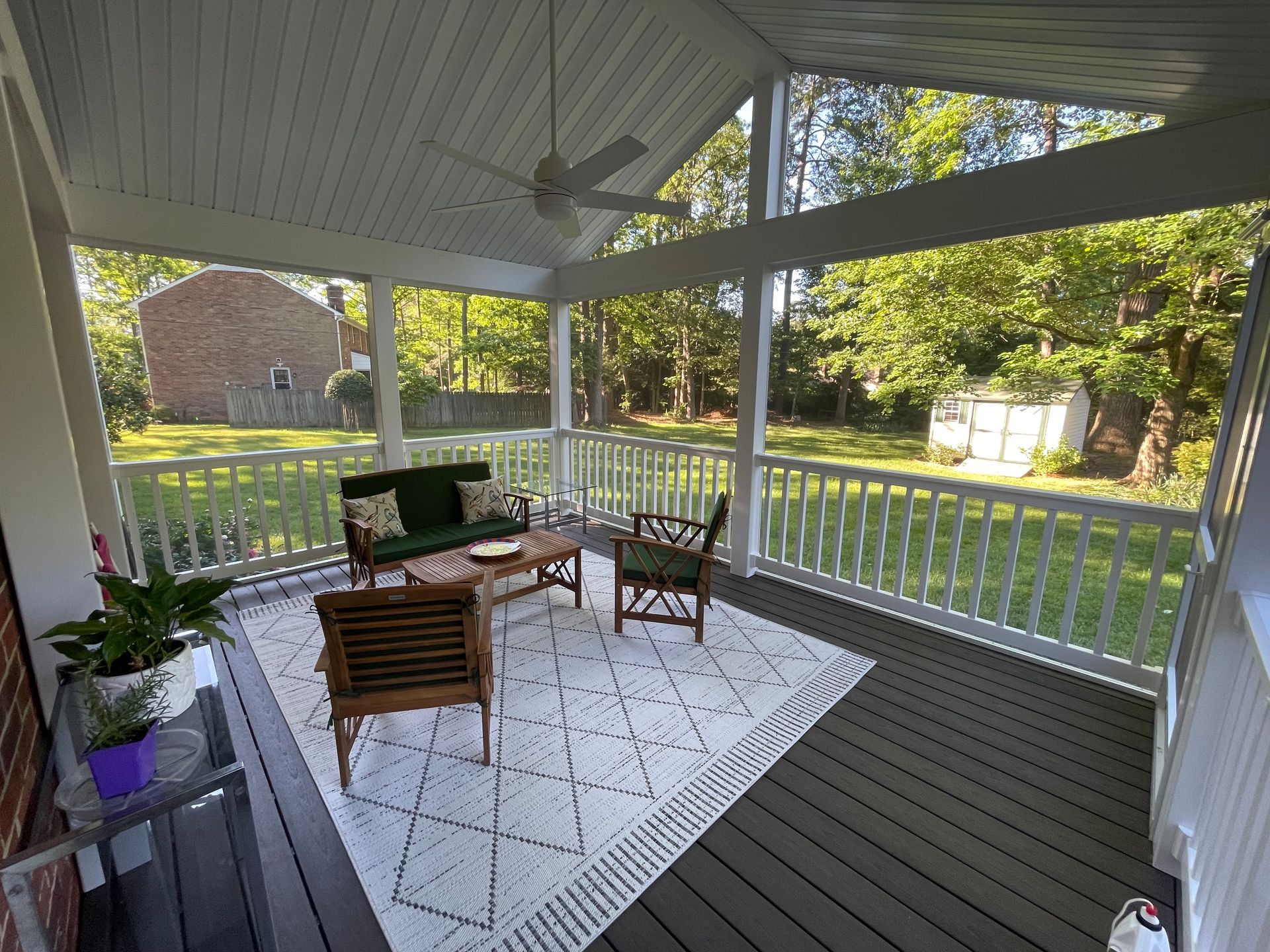 Covered porch with white railing, gray floor, white rug, wooden furniture, and potted plants overlooking a green yard