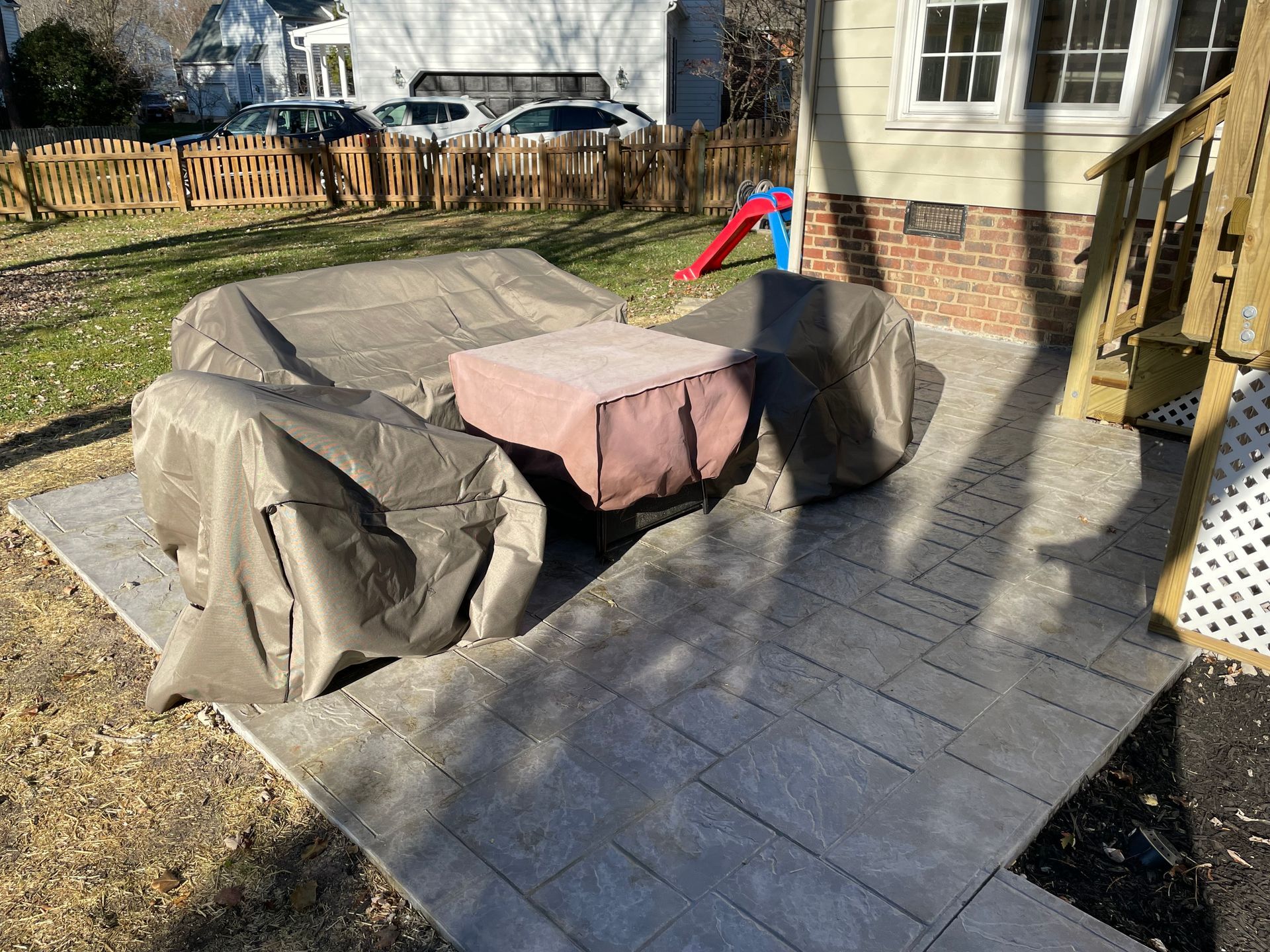 Covered patio furniture with a pink cushion on a gray stone patio beside a house and fenced yard
