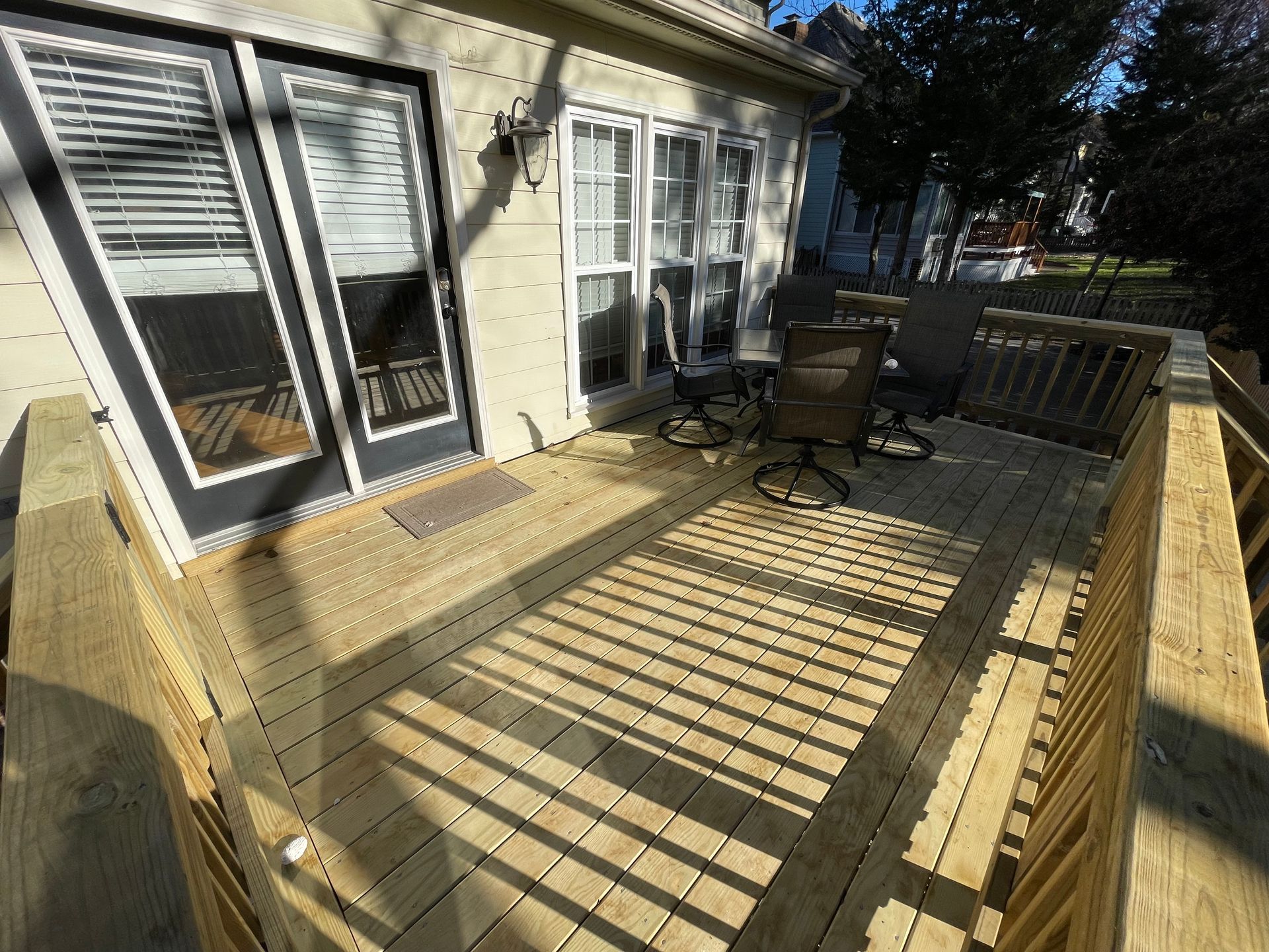 Backyard wooden deck with patio furniture beside house windows, lit by sun and shadows