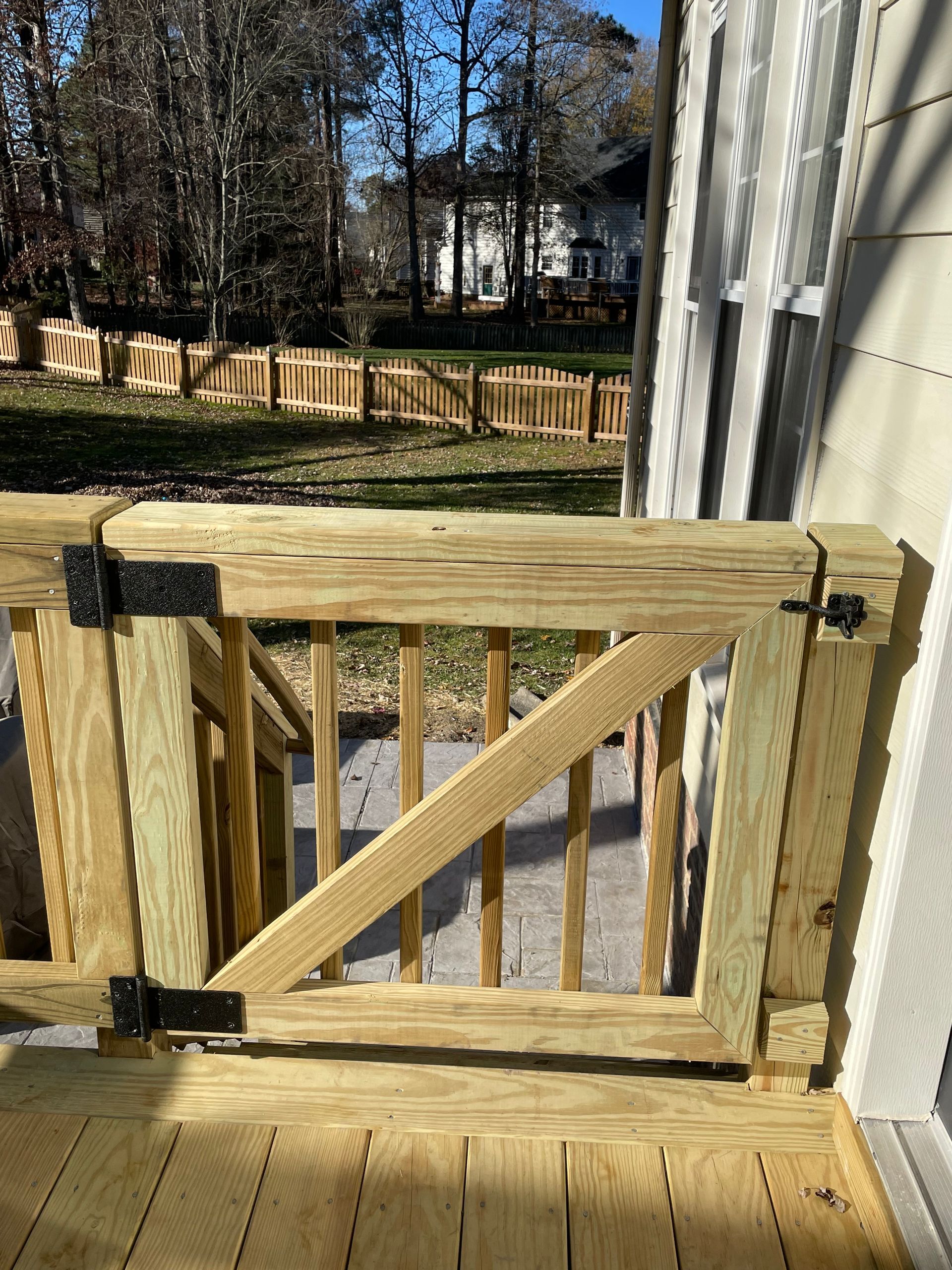 Wooden backyard gate with diagonal brace beside a house, opening to a fenced yard with trees beyond
