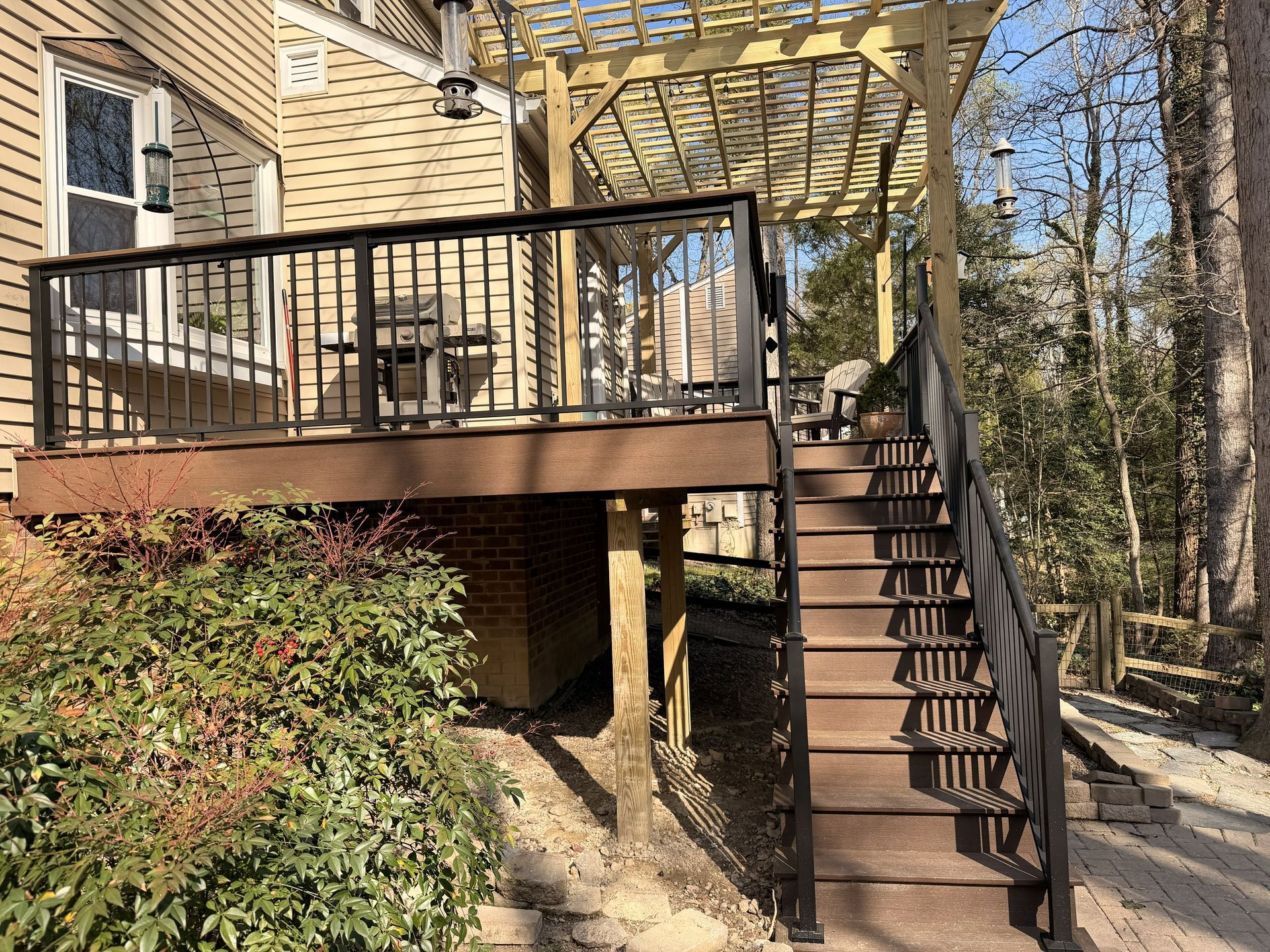 Raised wooden deck with black railing and stairs beside a beige house, surrounded by trees and shrubs
