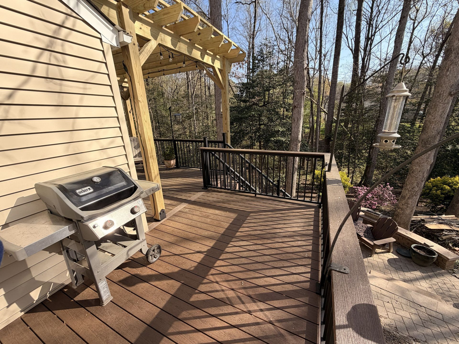 Wooden deck beside a house with a grill and black railing, overlooking a wooded backyard.