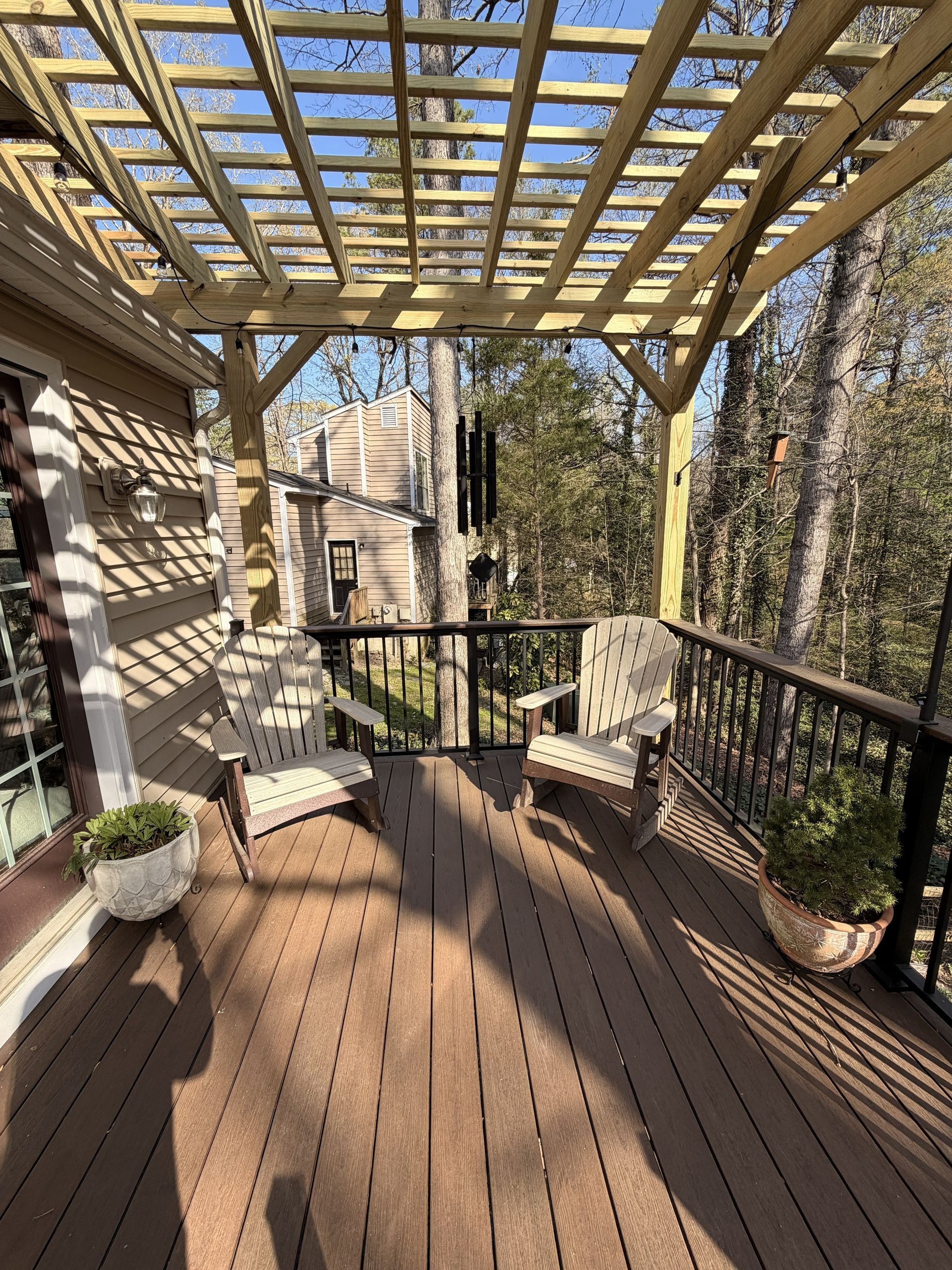 Wooden deck under a pergola, with chairs, potted plants, and a view of the wooded yard.