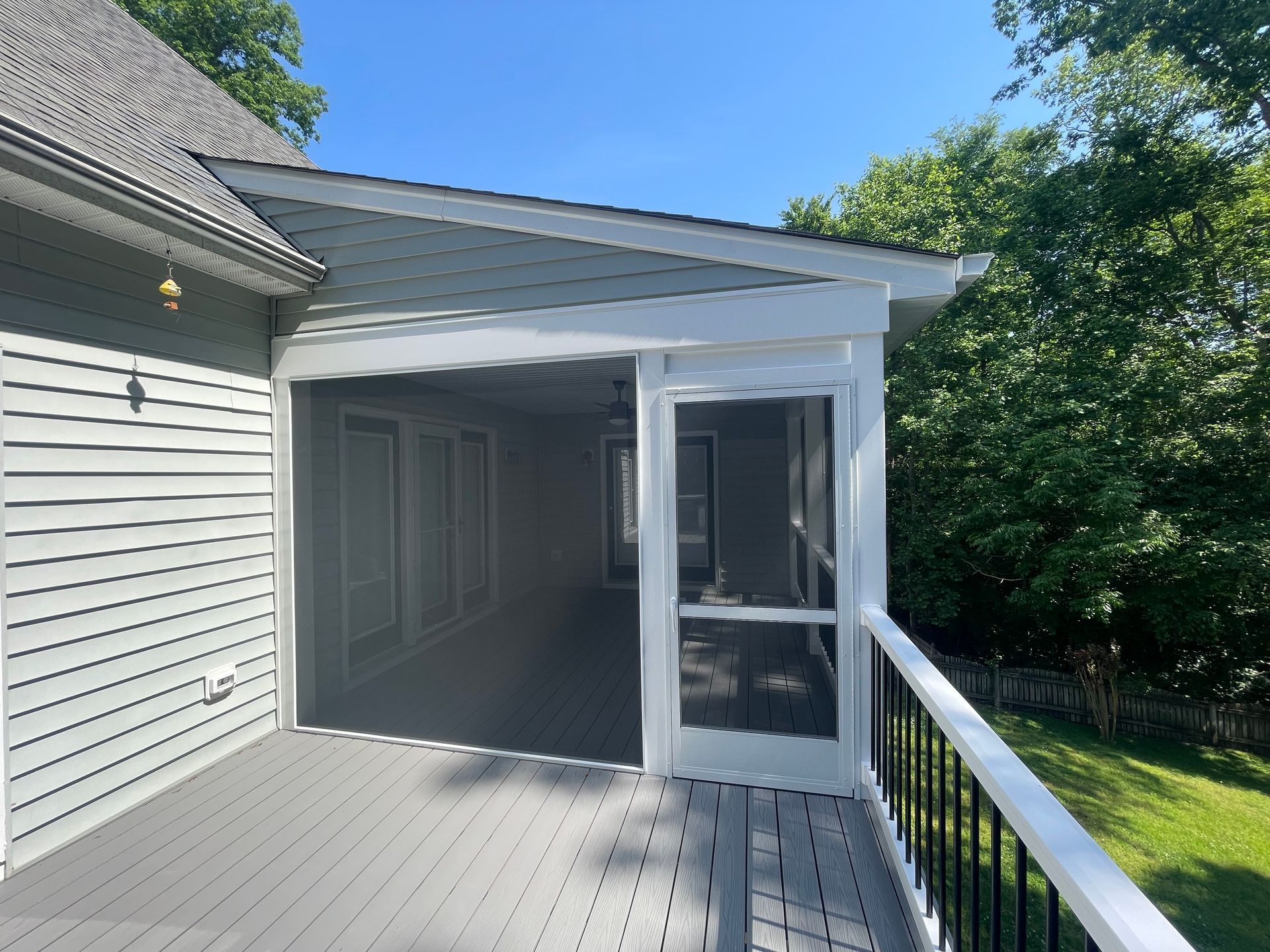 Covered gray deck with screened porch beside a white house, bordered by trees.